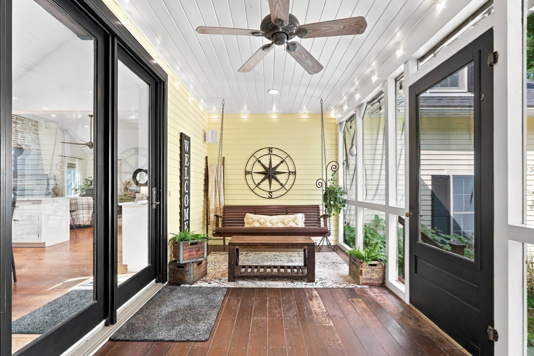 Enclosed sunroom with a wooden porch swing, compass wall decor, and black-framed glass doors.