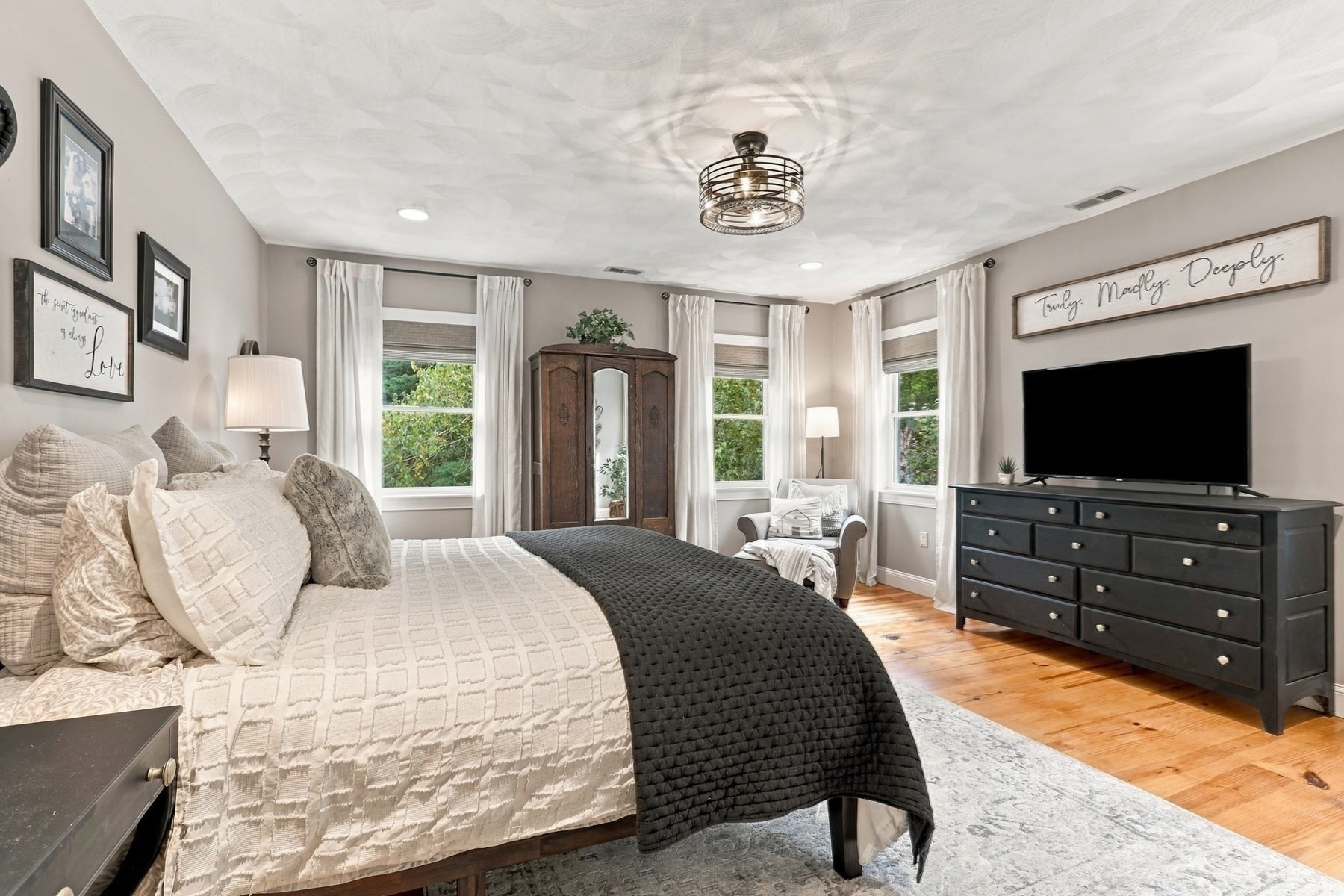 Staged bedroom with cream bedding, a dark quilt, hardwood floors, and three curtained windows.