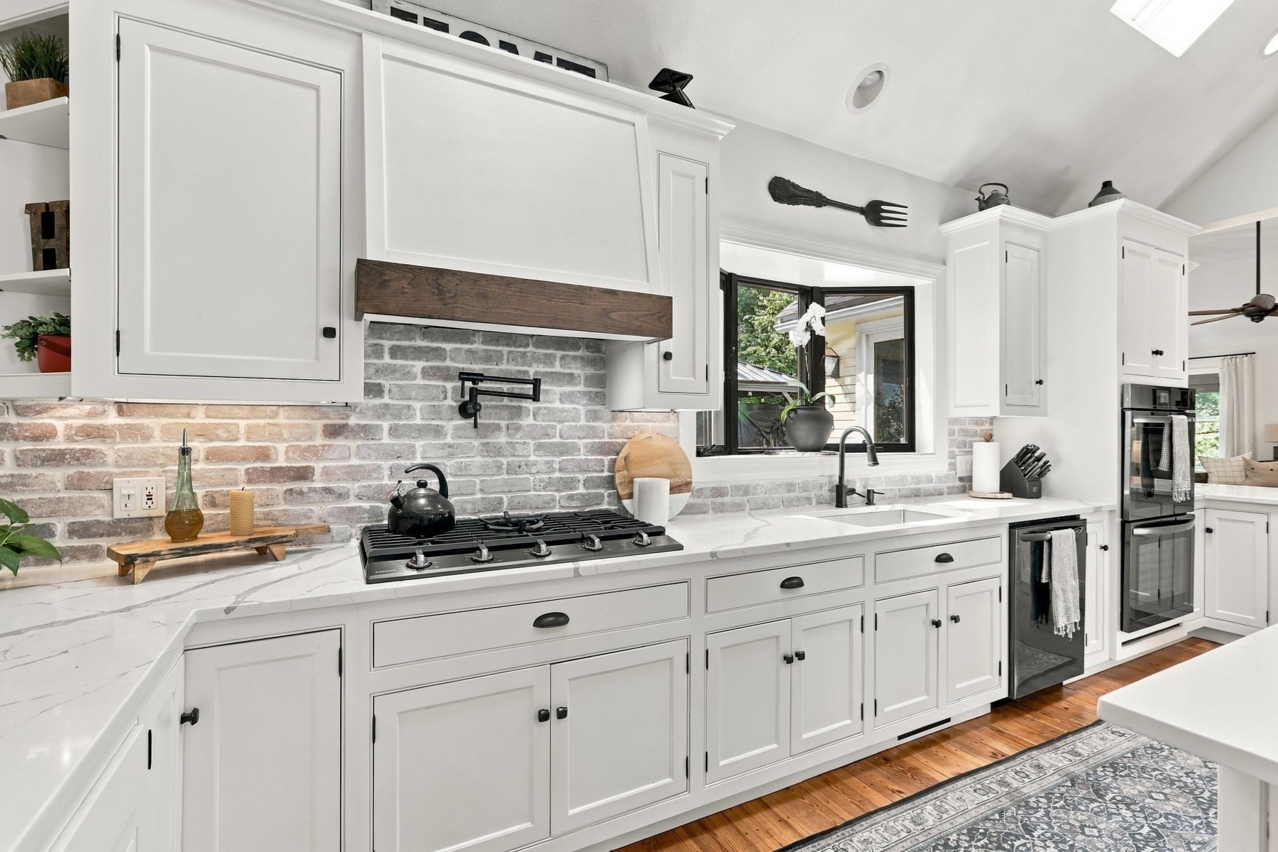 Bright kitchen with white cabinets, pale brick backsplash, black gas cooktop, and farmhouse sink beneath a wide window.