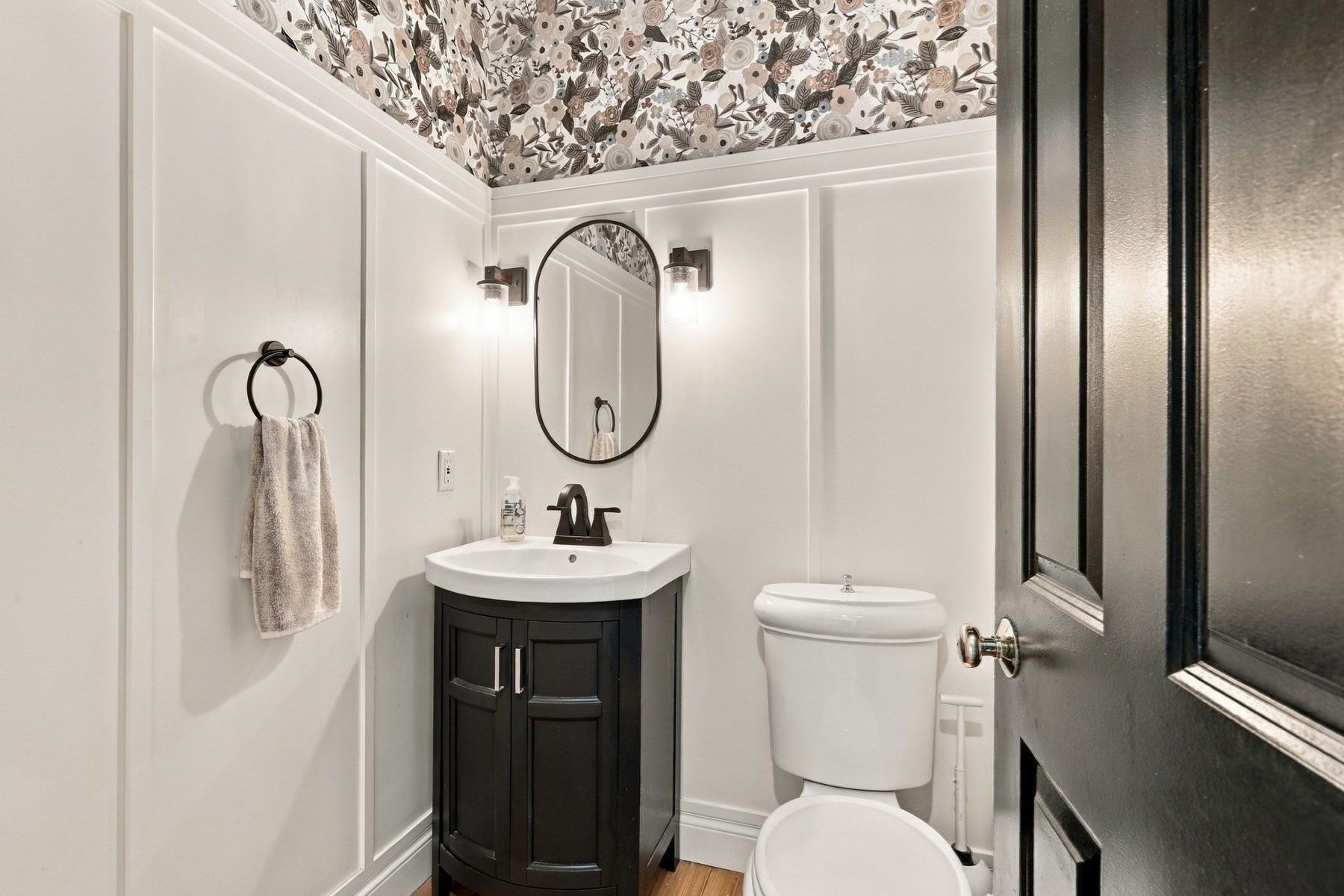 Powder room with a dark vanity, oval mirror, toilet, and floral ceiling wallpaper.