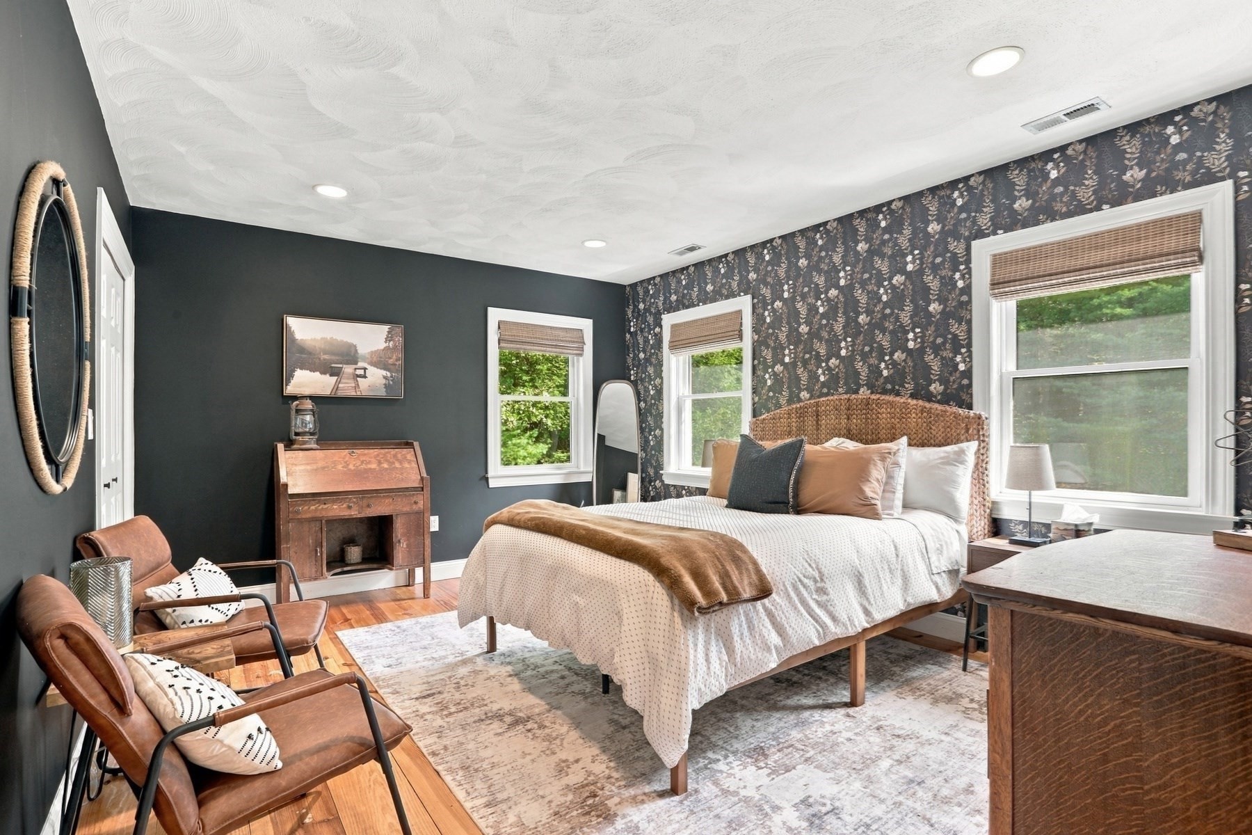 Bedroom with a woven-headboard bed, floral wallpaper, dark teal wall, and leather chairs.