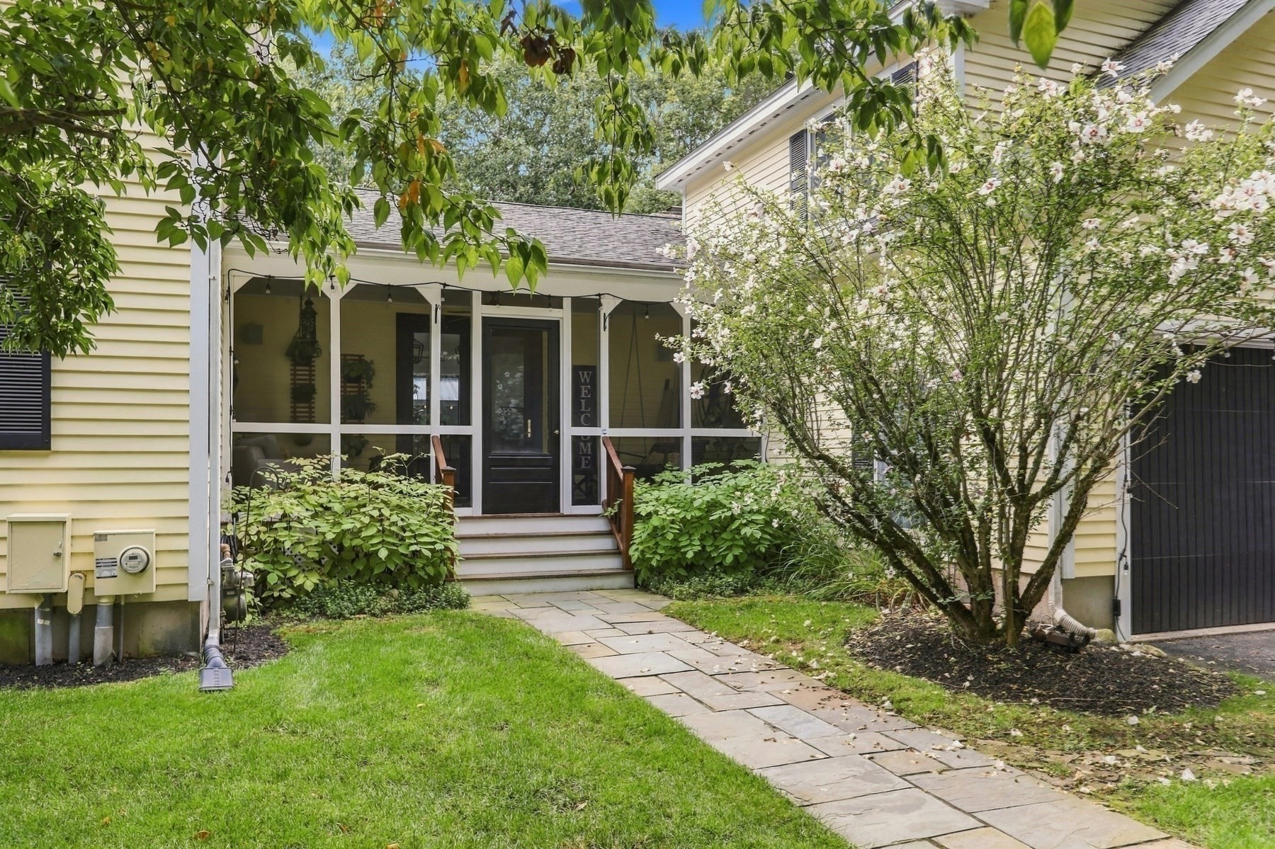 Yellow-sided home with a screened porch, stone walkway, and flowering shrubs.