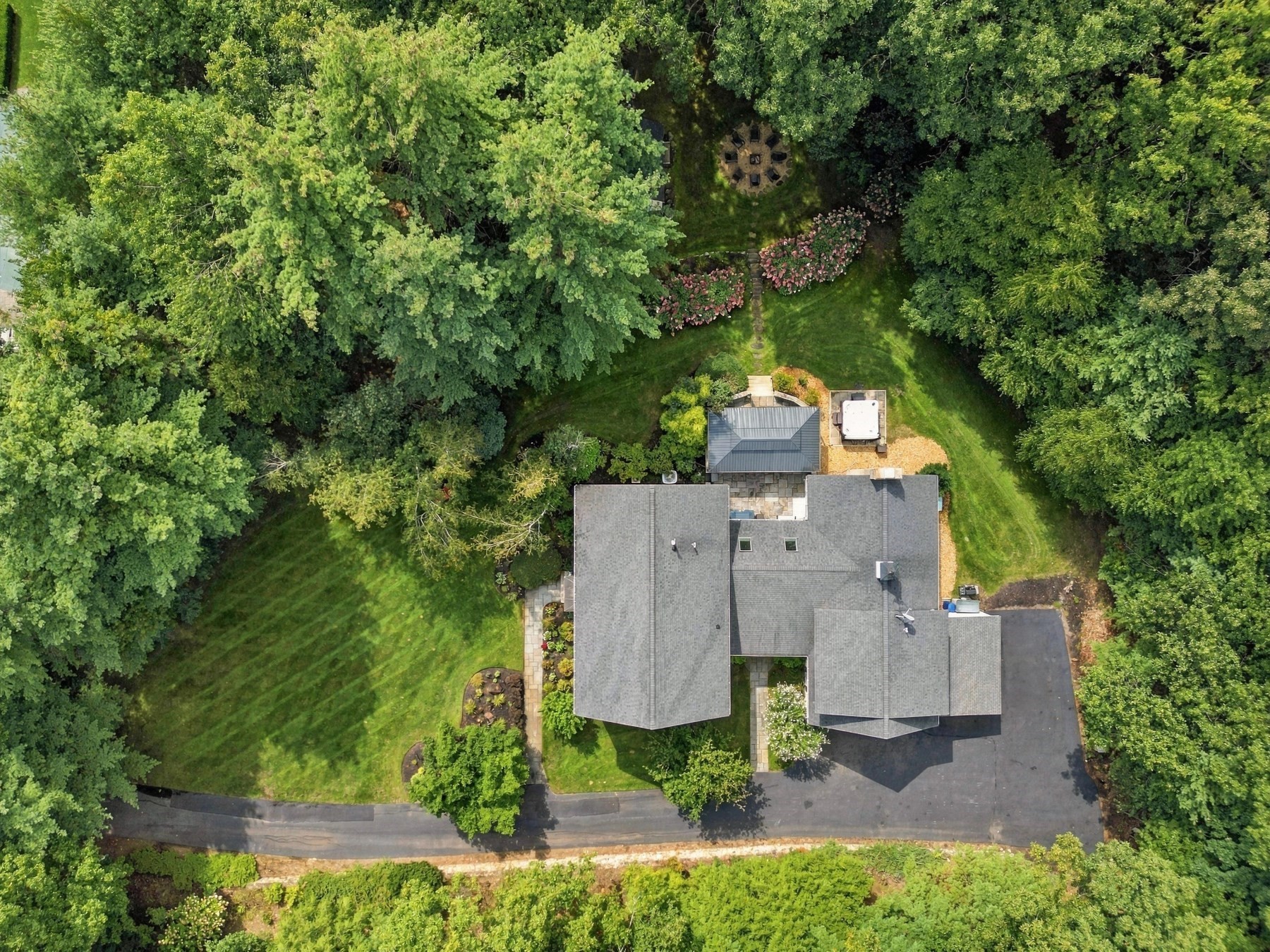 Aerial view of a house with a wooded lawn, stone patio, hot tub, and circular fire pit.
