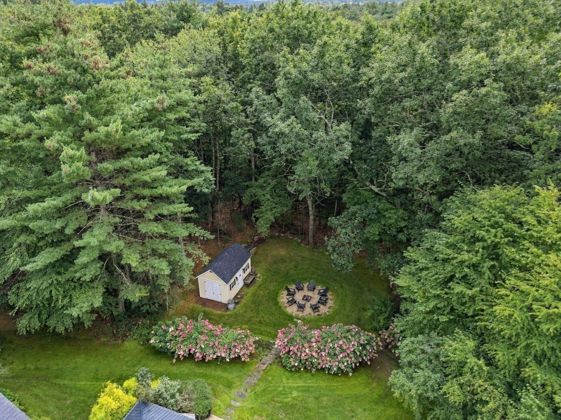 Aerial view of a wooded backyard with a shed, fire pit seating area, and pink flowering shrubs.