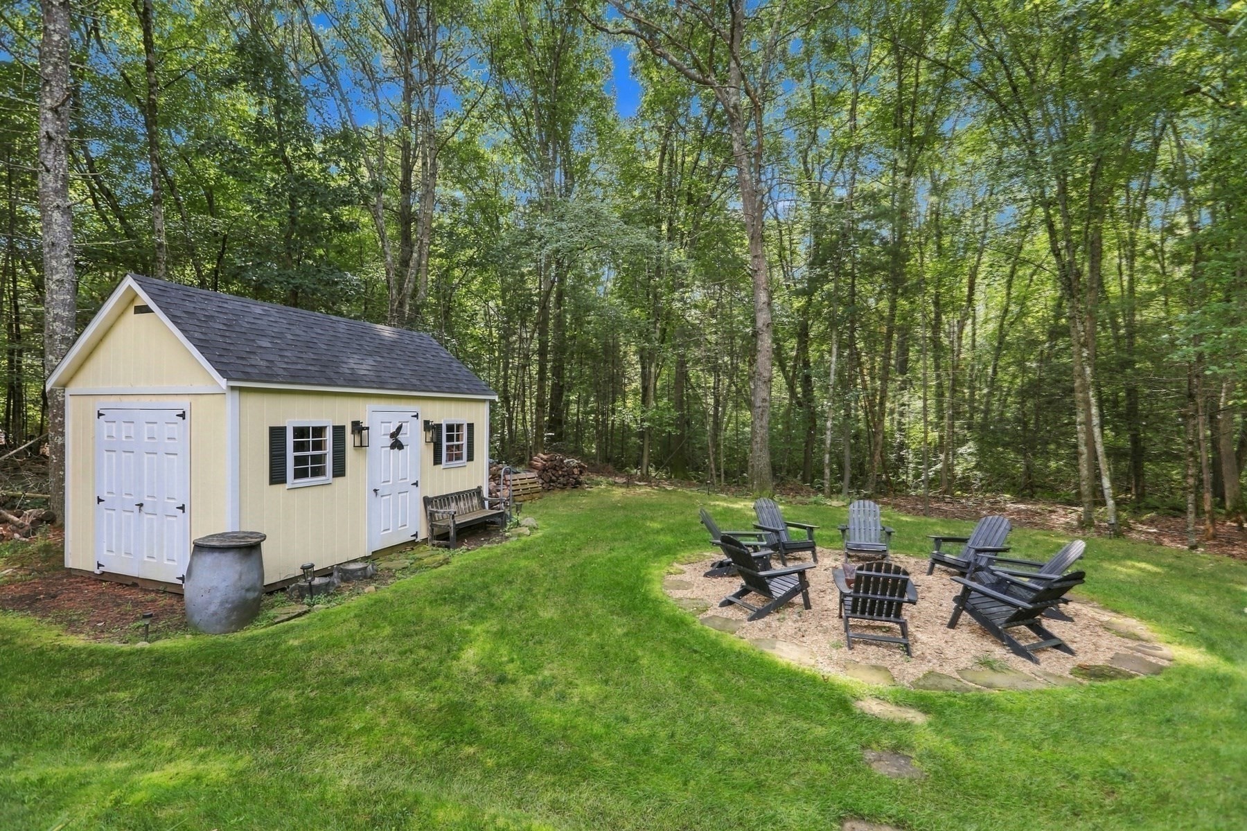 Backyard with a pale yellow shed, Adirondack chairs around a stone fire pit, and surrounding woods.