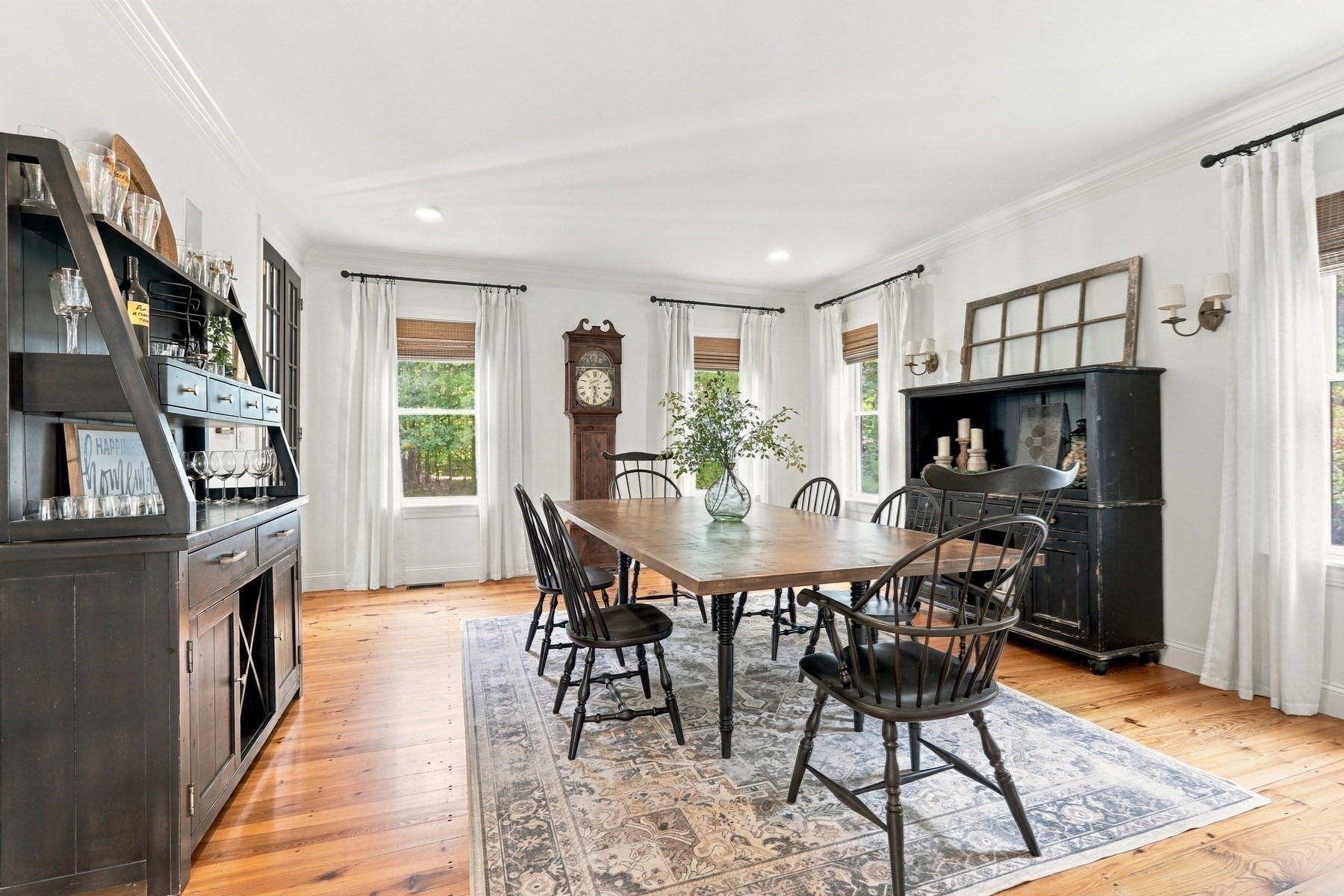 Dining room with a long wooden table, patterned rug, grandfather clock, and dark display cabinets.