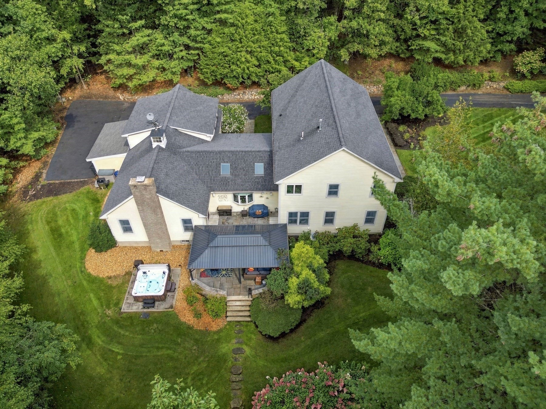 Aerial view of a cream-colored house with a wooded lawn, covered patio, and outdoor hot tub.