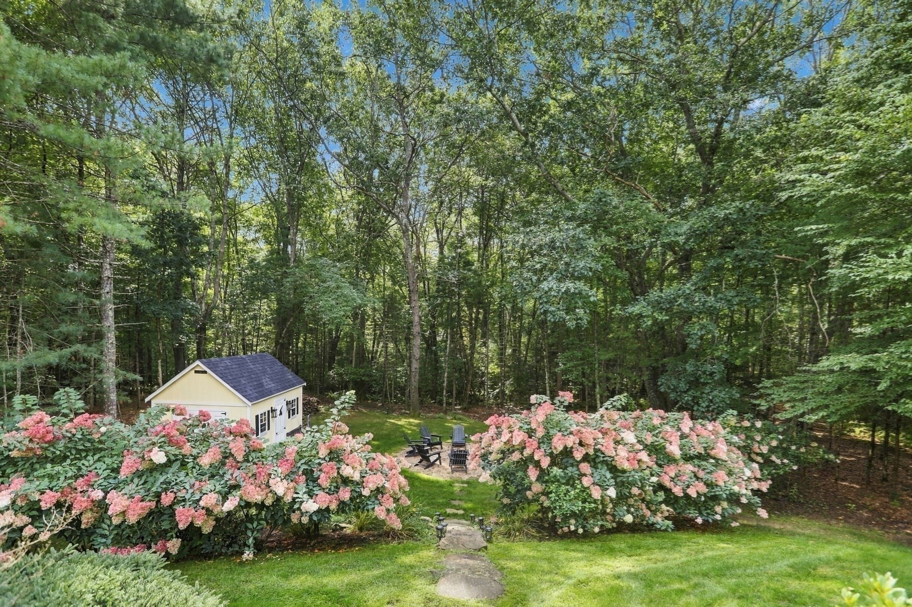 Wooded backyard with pink flowering shrubs, a yellow shed, and chairs around a fire pit.
