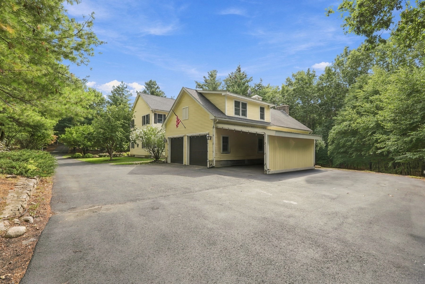 Yellow house with an attached garage and covered carport beside a broad driveway, surrounded by mature trees.