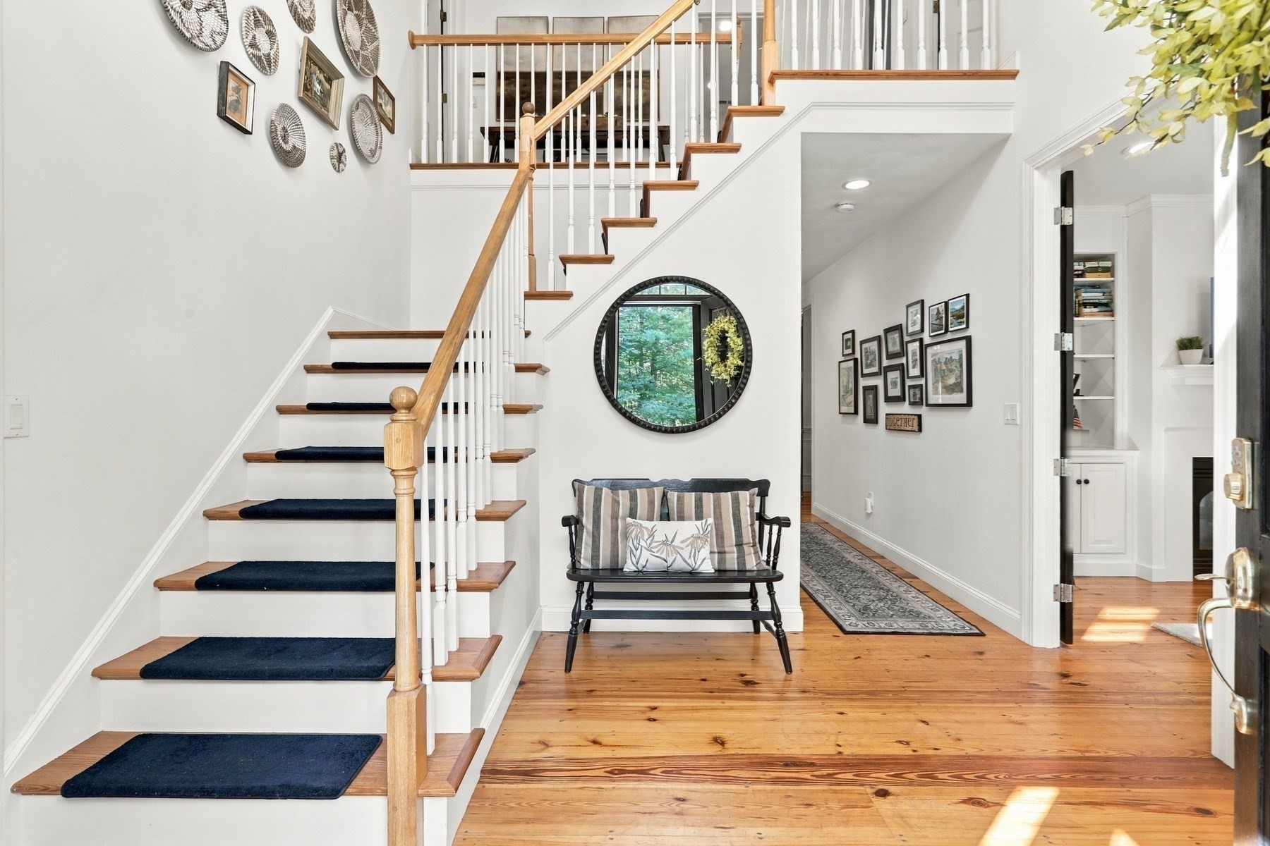 Home entryway with a wood staircase, navy runners, plank flooring, and a bench beneath a round mirror.