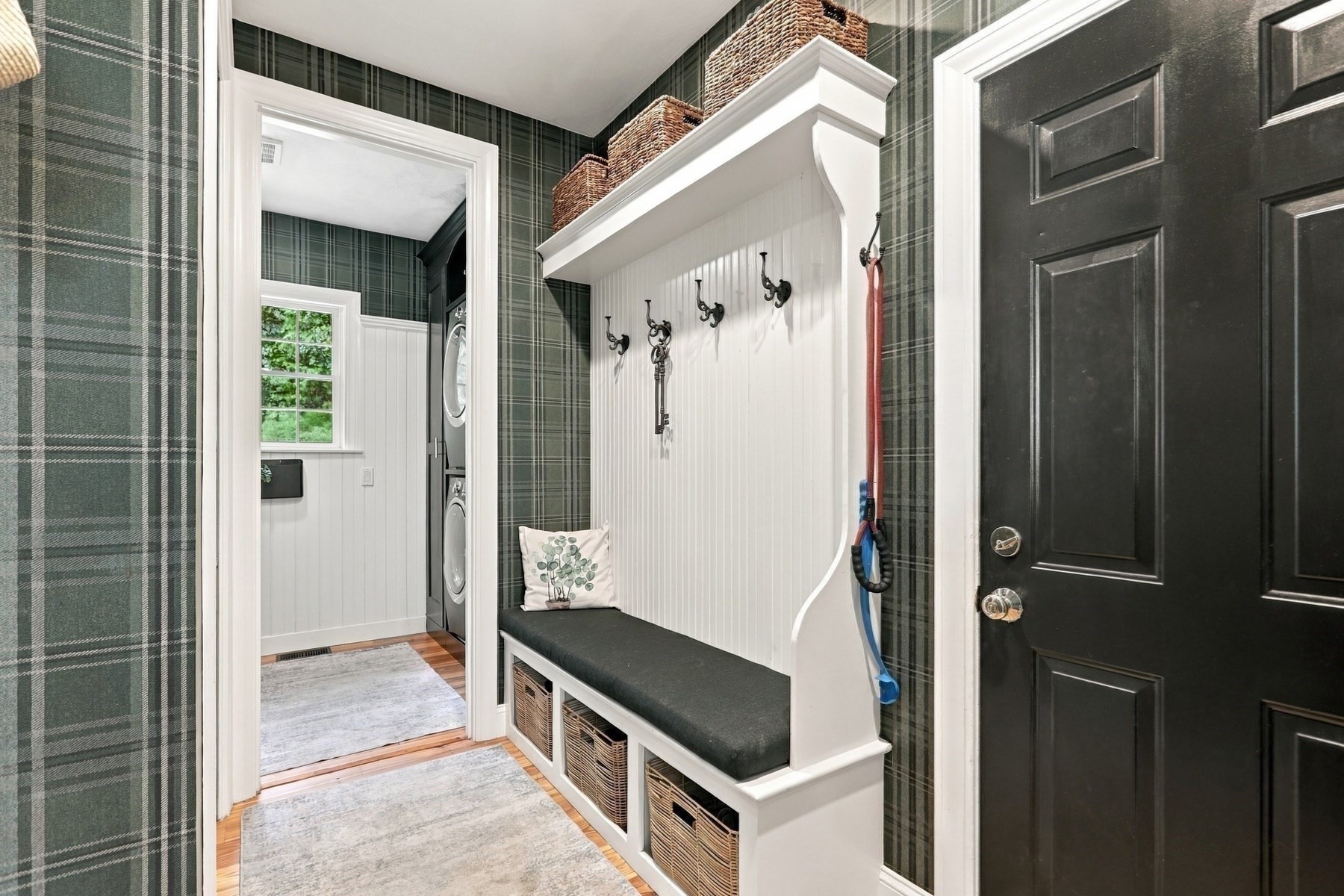 Mudroom with a built-in bench, coat hooks, wicker baskets, plaid wallpaper, and a laundry area beyond.