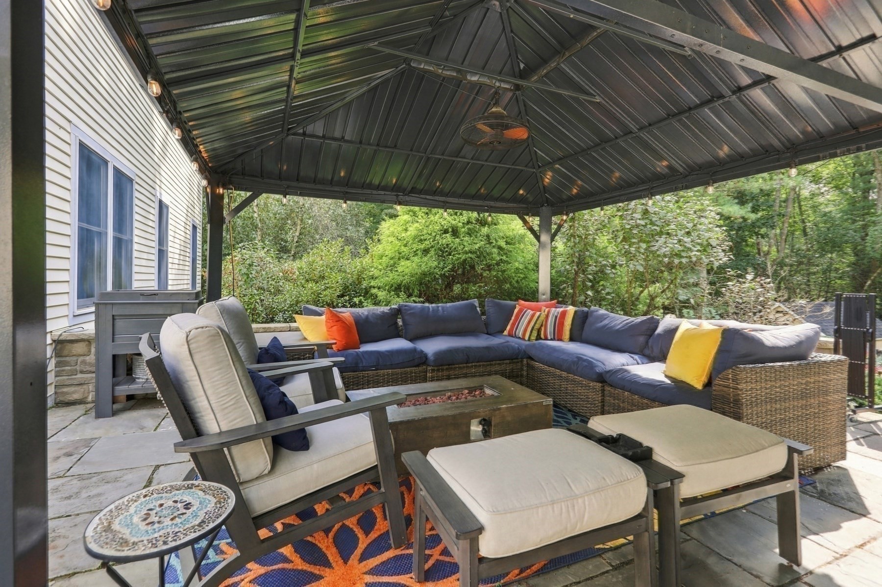 Covered patio with sectional seating and a fire table, surrounded by greenery.