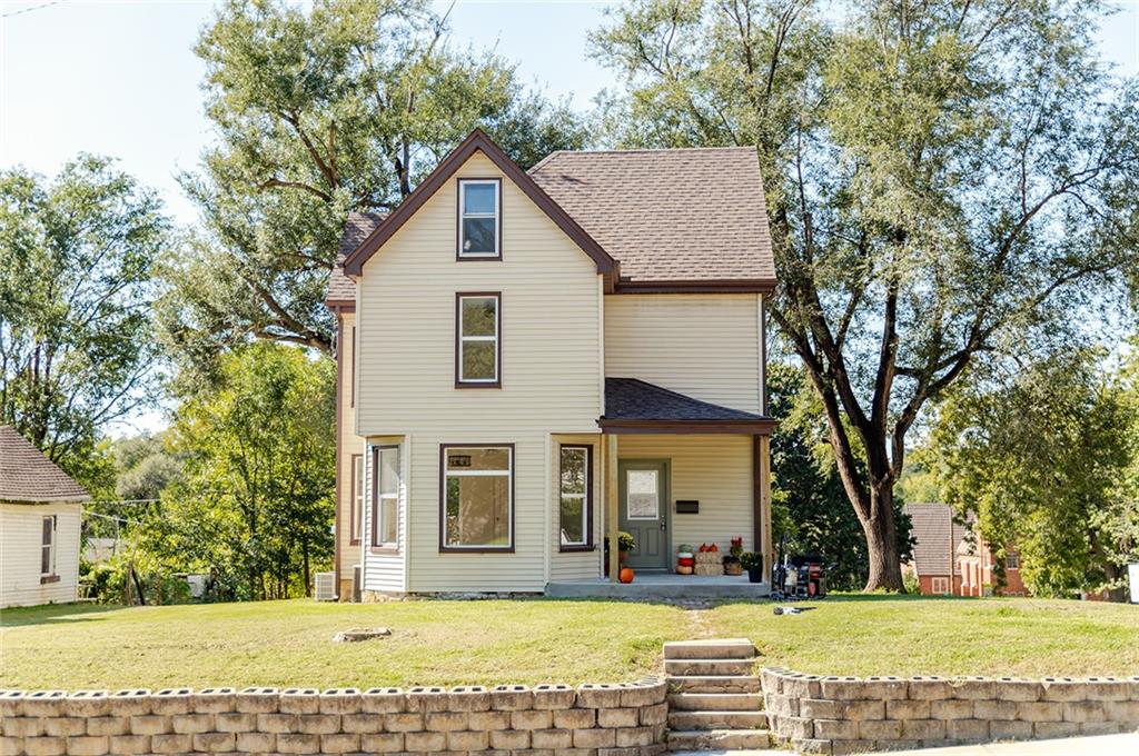 Two-story cream house with a covered entry, mature trees, and a stone retaining wall.