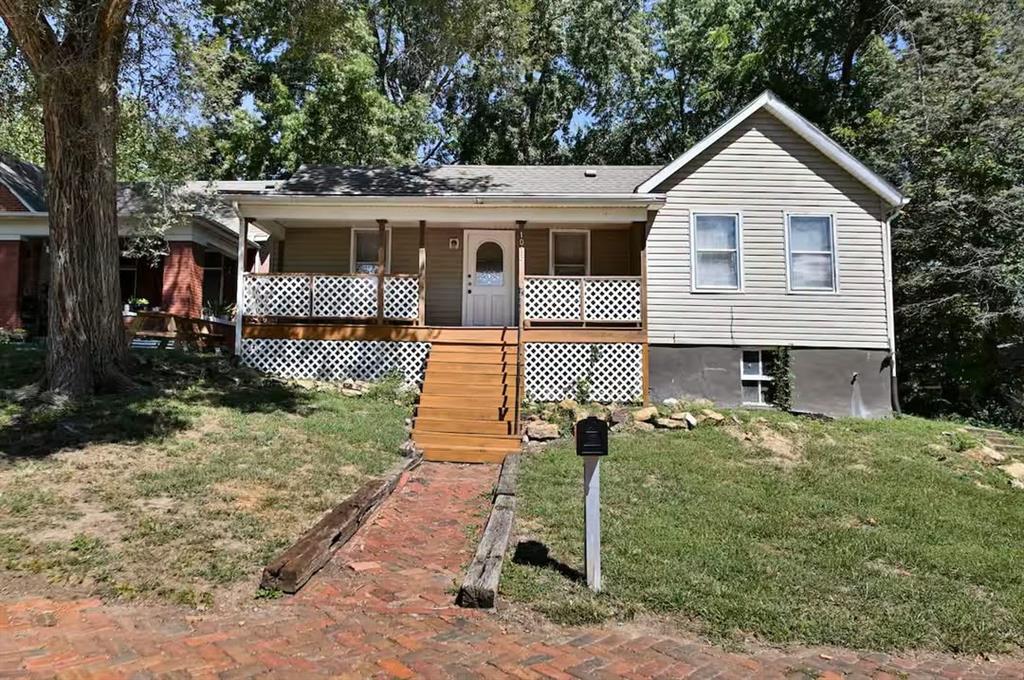 Light-sided house with a lattice-front porch, wooden steps, and a brick walkway through a tree-shaded yard.