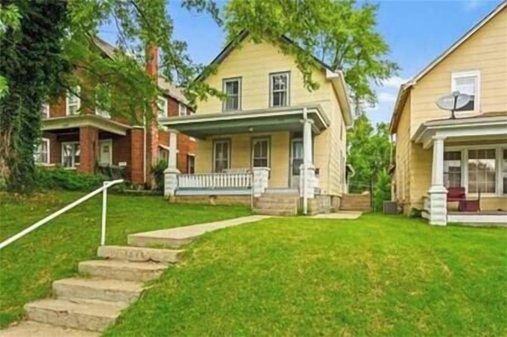 Yellow two-story house with a covered porch, white railing, and stone steps across a green lawn.