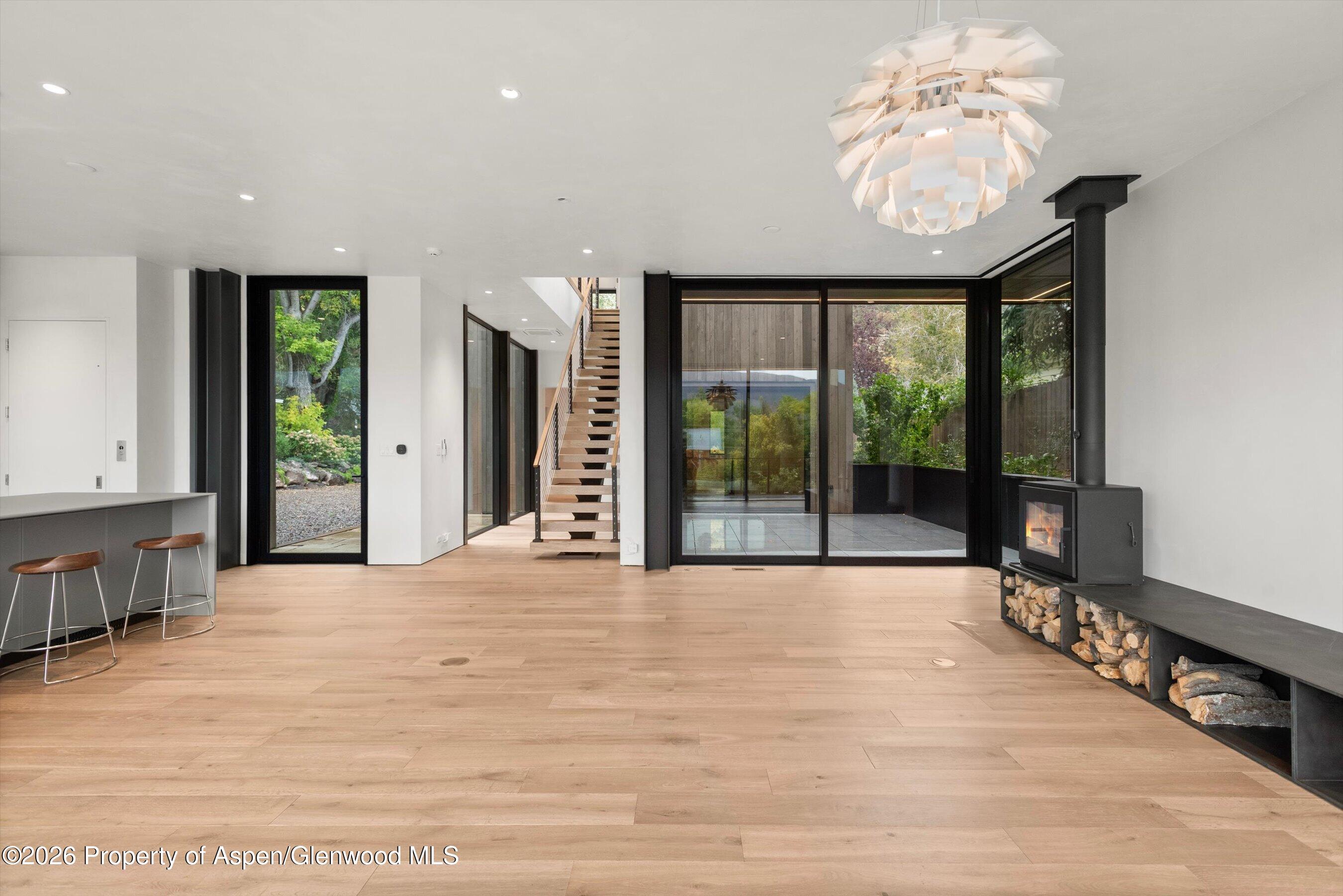 Modern open-plan room with wood floors, an open staircase, glass patio doors, and a wood-burning stove.