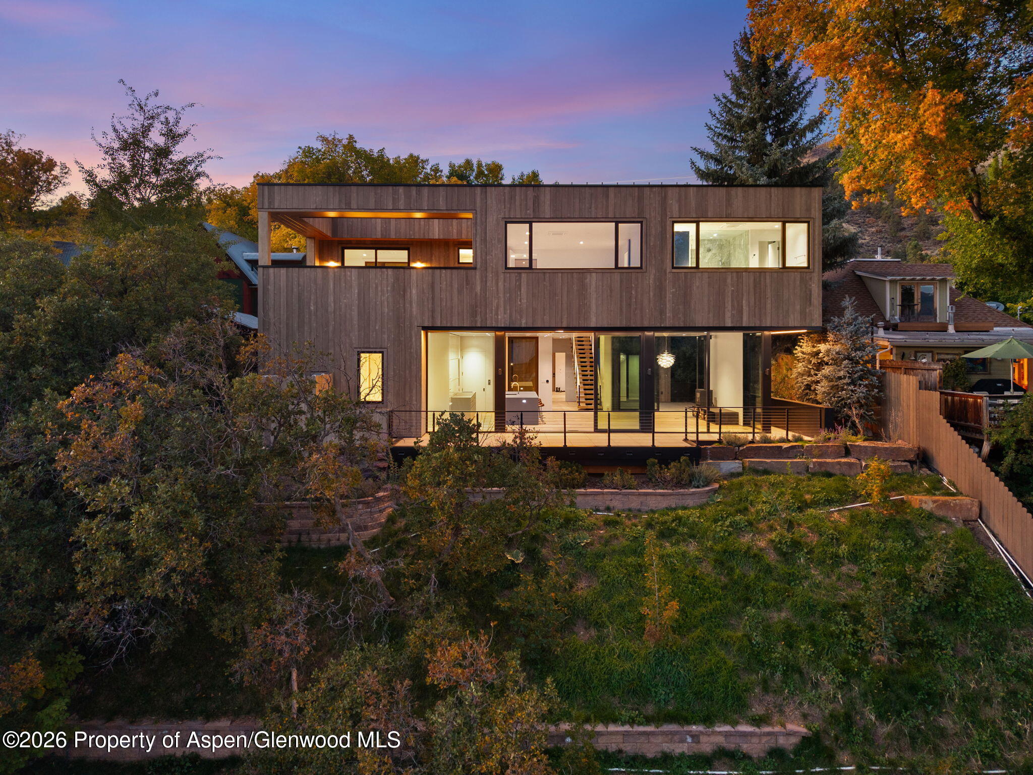 Modern wood-clad home with illuminated windows and a deck on a landscaped hillside at dusk.