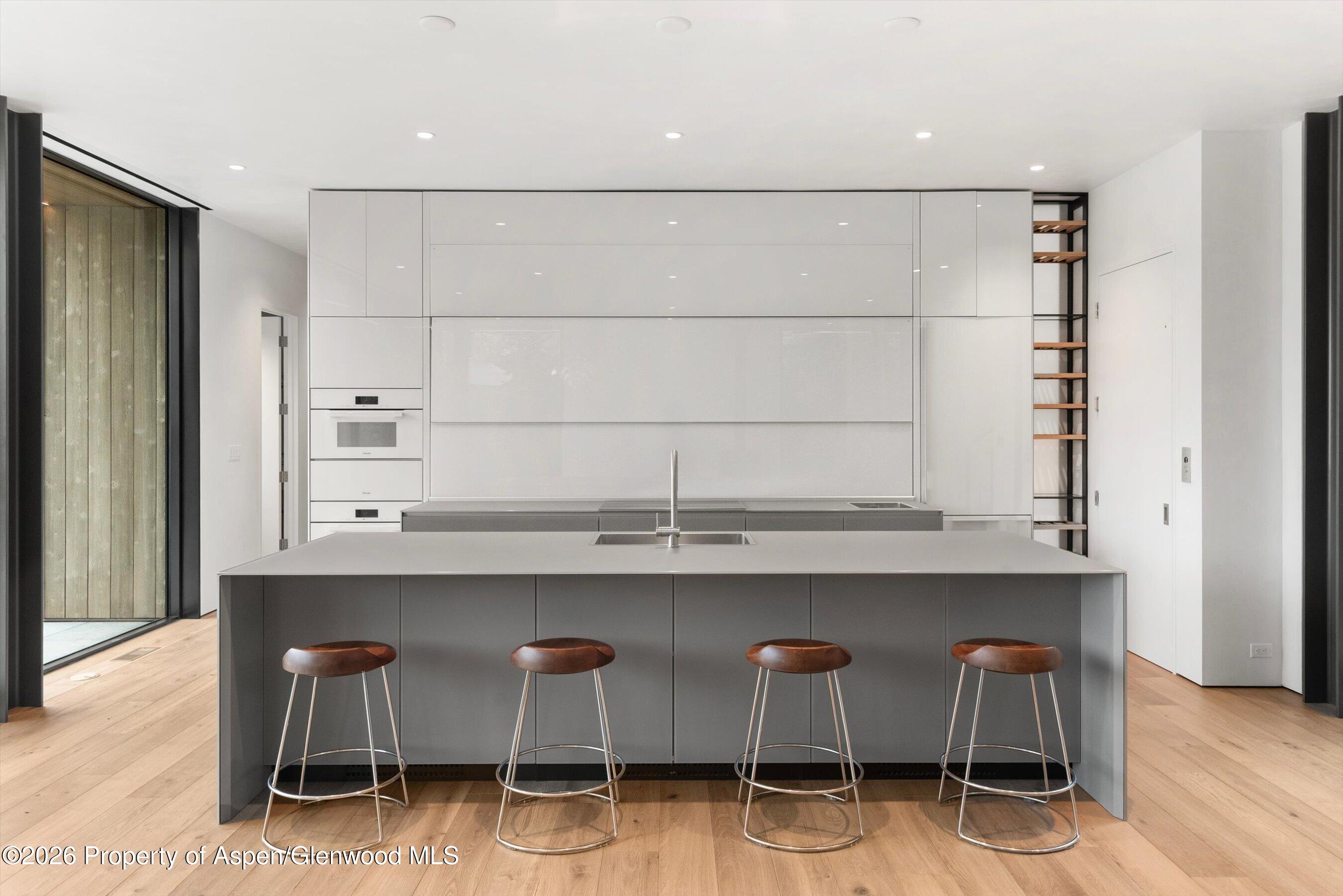 Modern kitchen with a large gray island, four bar stools, flush cabinetry, and light wood floors.