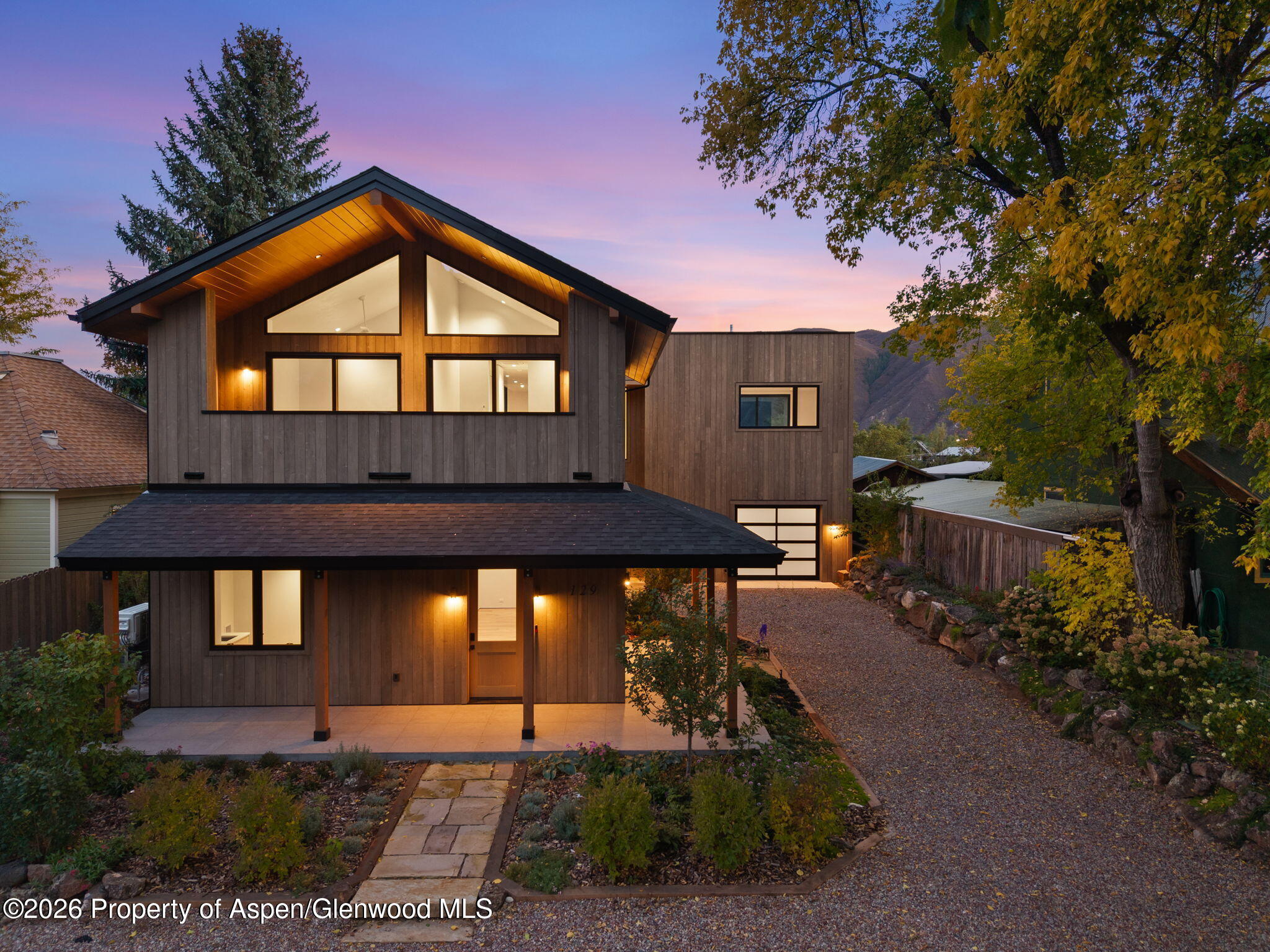 Modern wood-sided home at dusk with a steep gabled roof, warmly lit windows, and autumn trees.