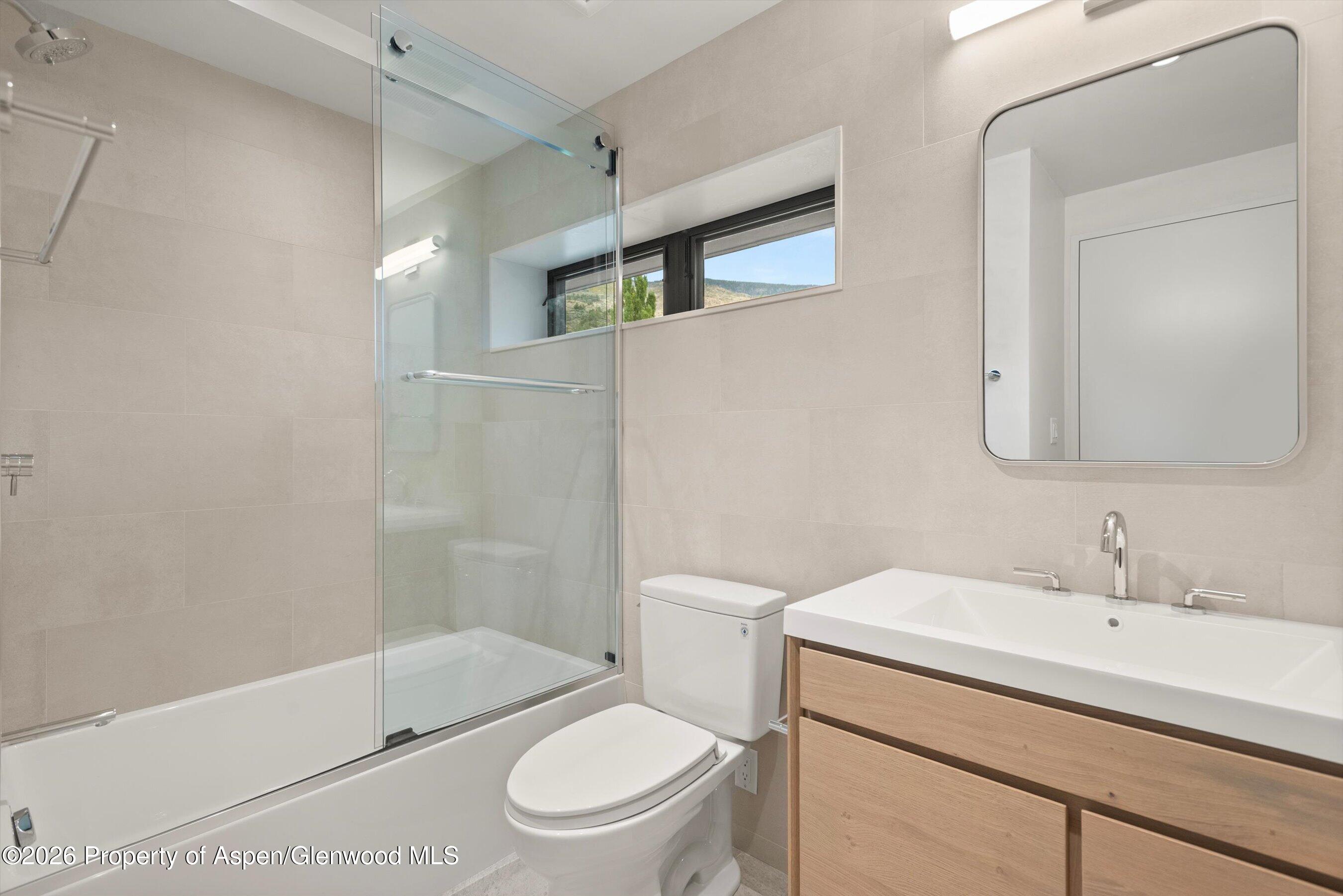 Modern bathroom with a glass-enclosed tub and shower, wood vanity, and narrow hillside-view window.