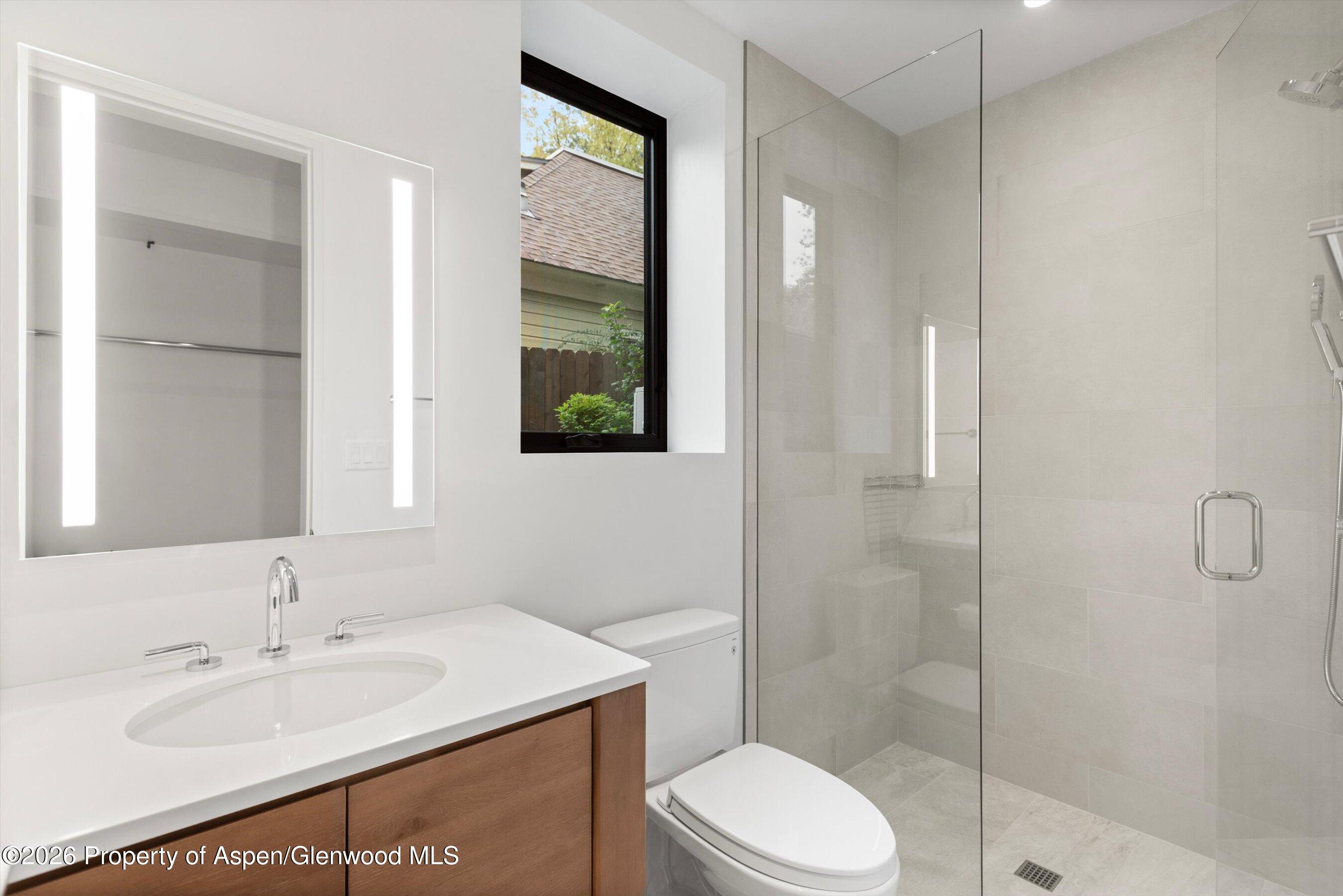 Modern bathroom with wood vanity, illuminated mirror, black-framed window, and glass-enclosed tiled shower.