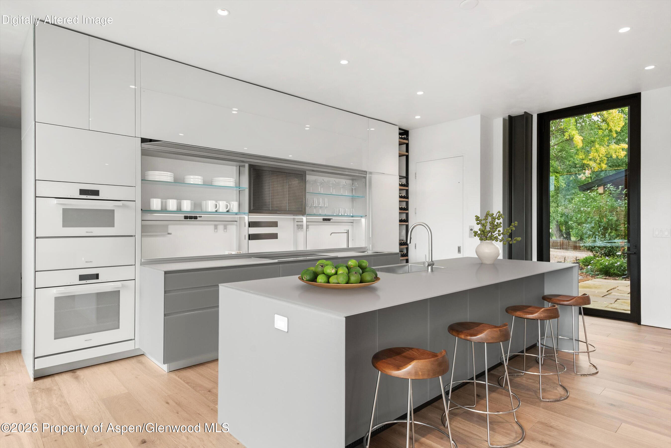 Modern kitchen with a large gray island, wooden bar stools, glossy cabinetry, and a garden view.