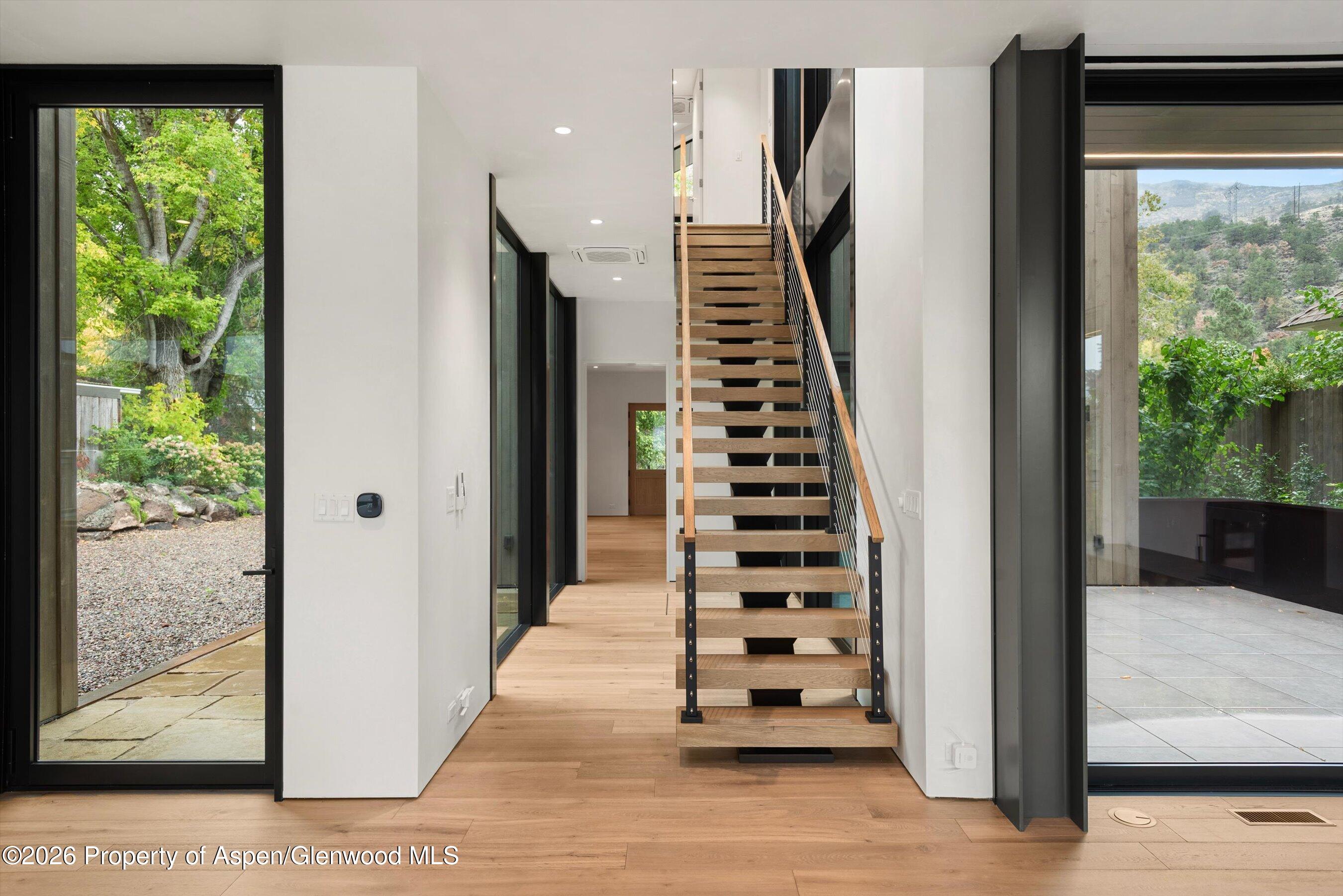 Modern entry hall with an open-riser wood staircase and glass doors overlooking greenery and mountains.