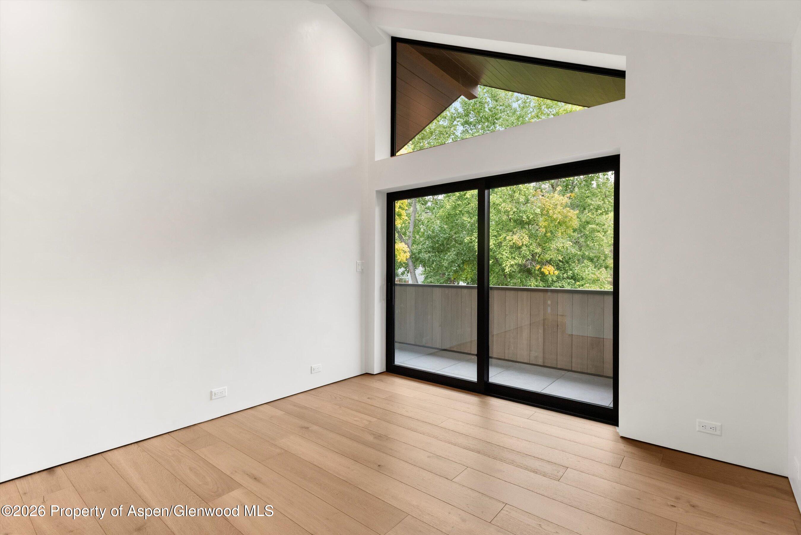 Empty modern room with wood floors, sliding glass doors, and a balcony overlooking trees.
