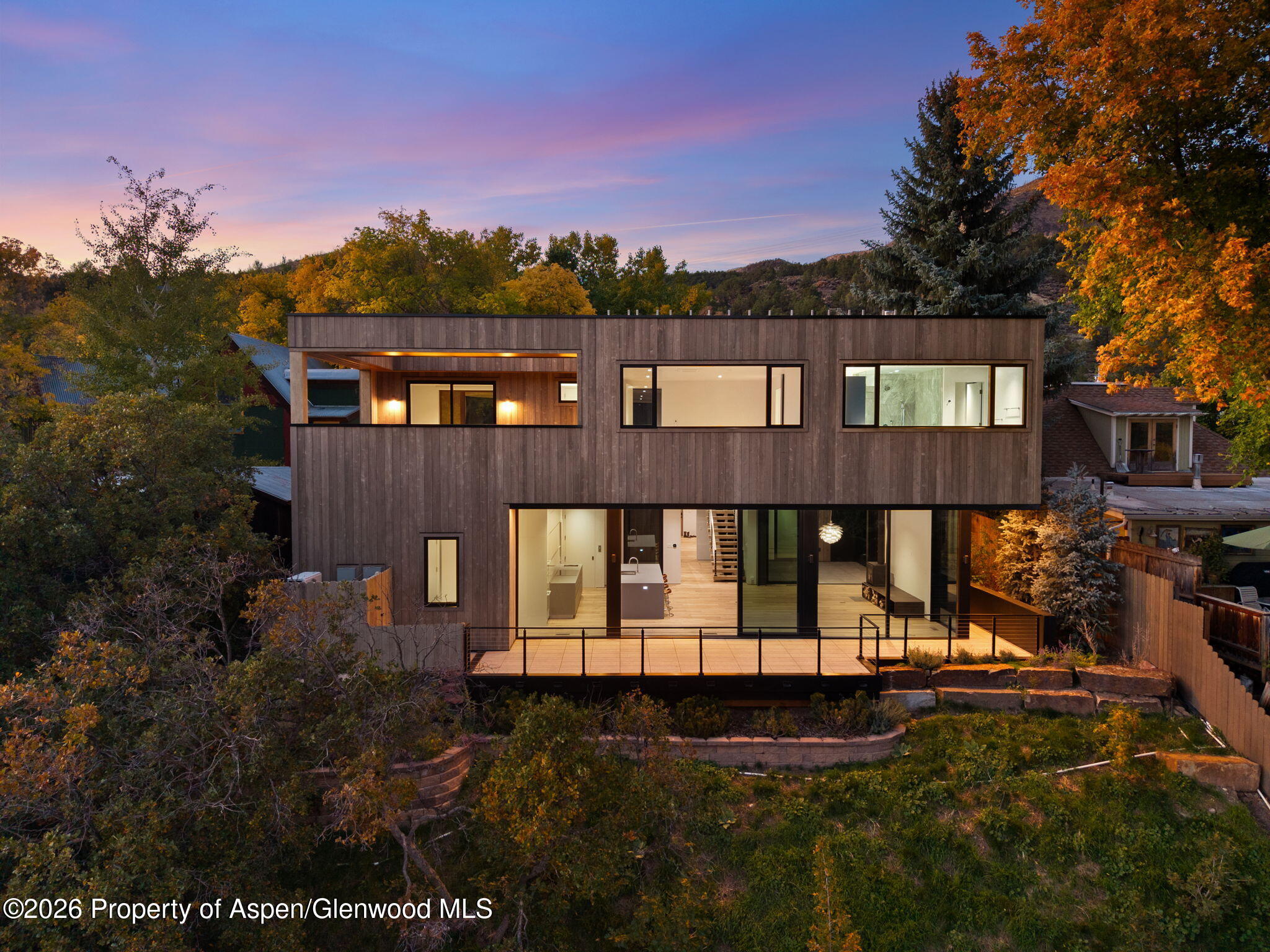 Modern two-story home with dark wood siding, broad windows, and an illuminated deck at dusk.