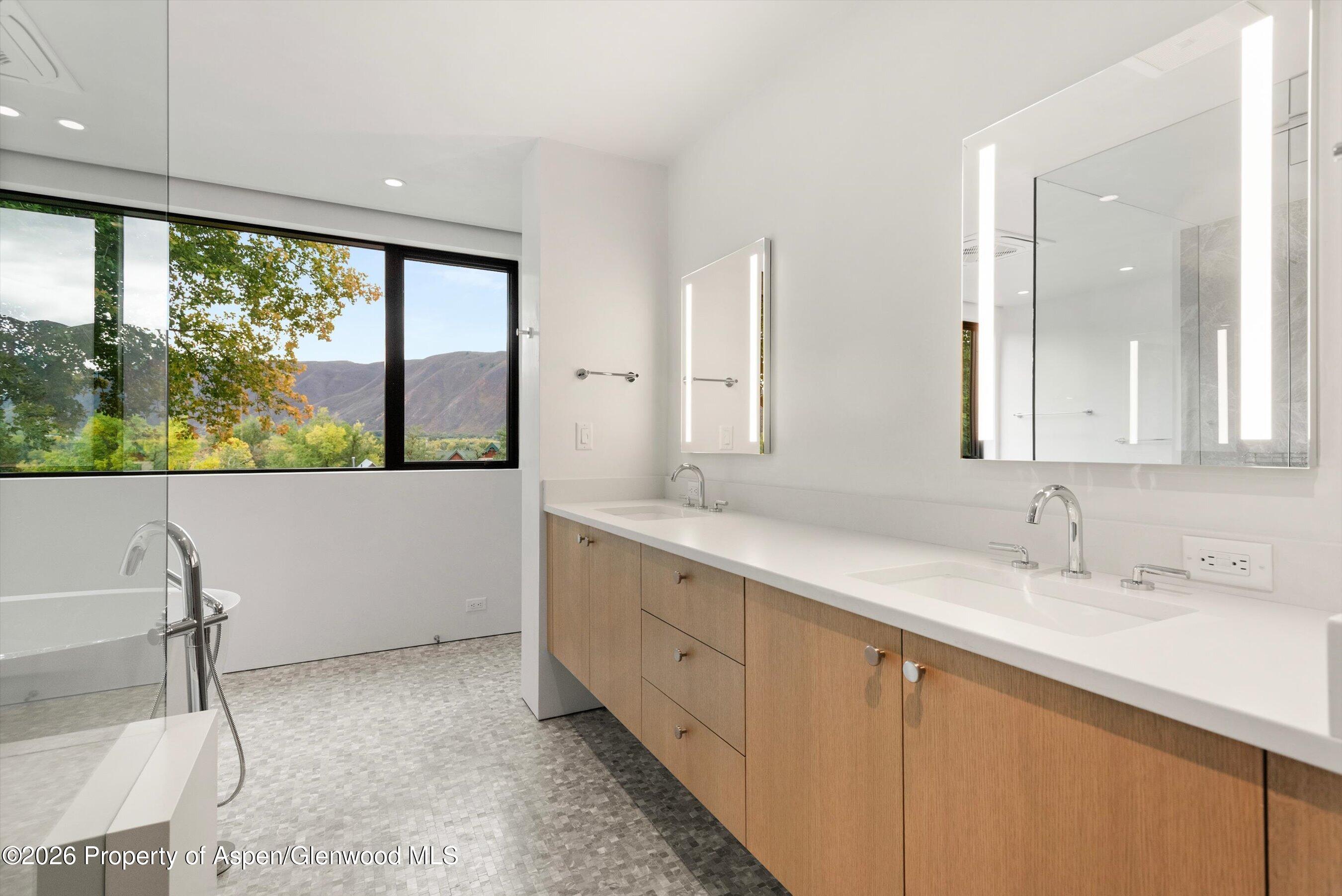 Modern bathroom with a wood double vanity and a large window overlooking autumn trees and mountains.