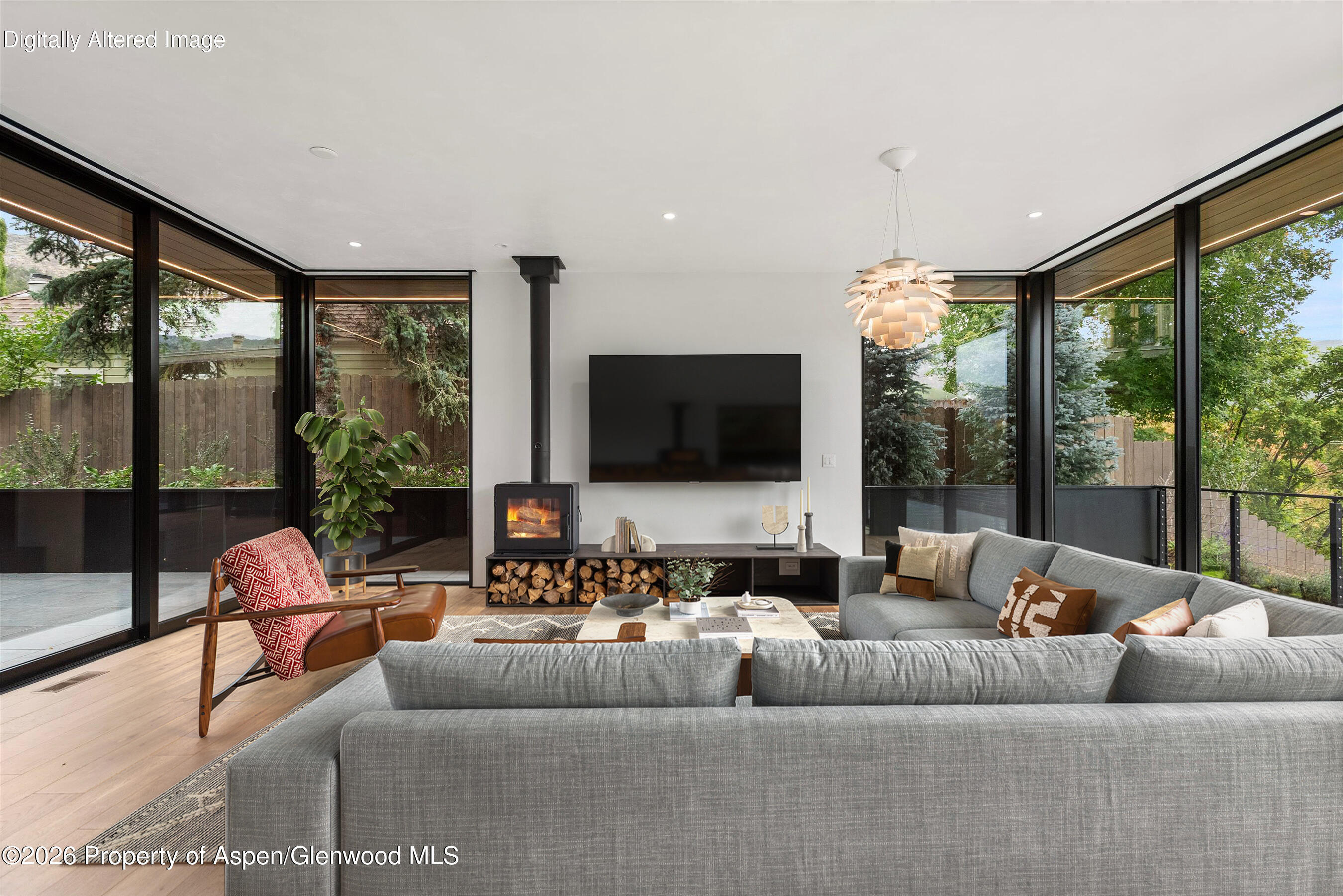 Furnished living room with wraparound windows, gray sectional, and a lit wood stove.