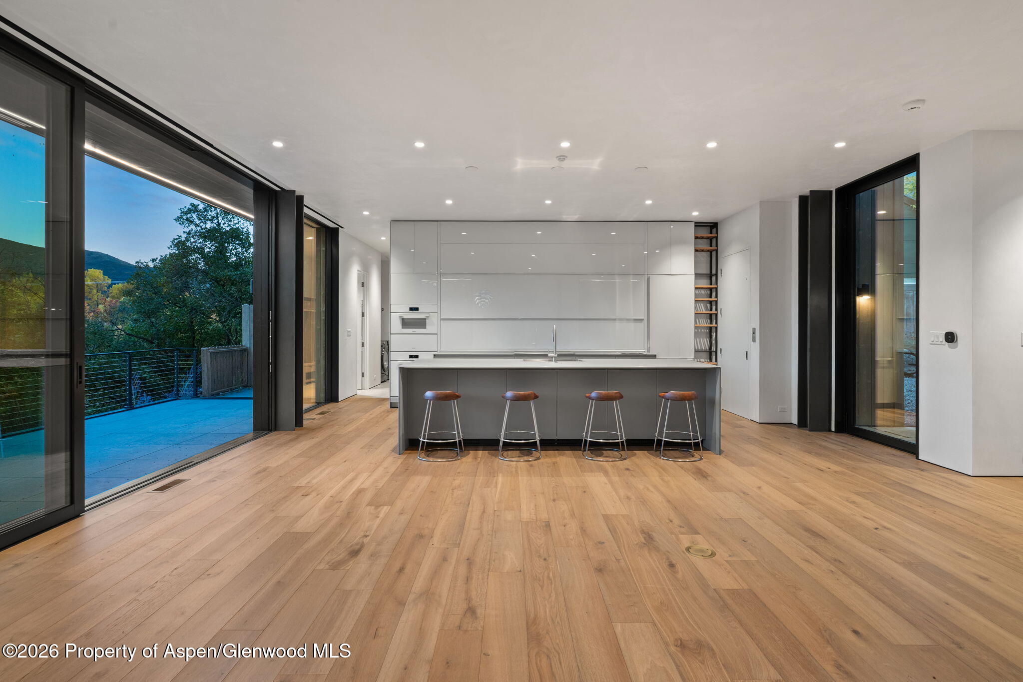 Modern kitchen with a gray island and four stools beside floor-to-ceiling doors opening to a deck.