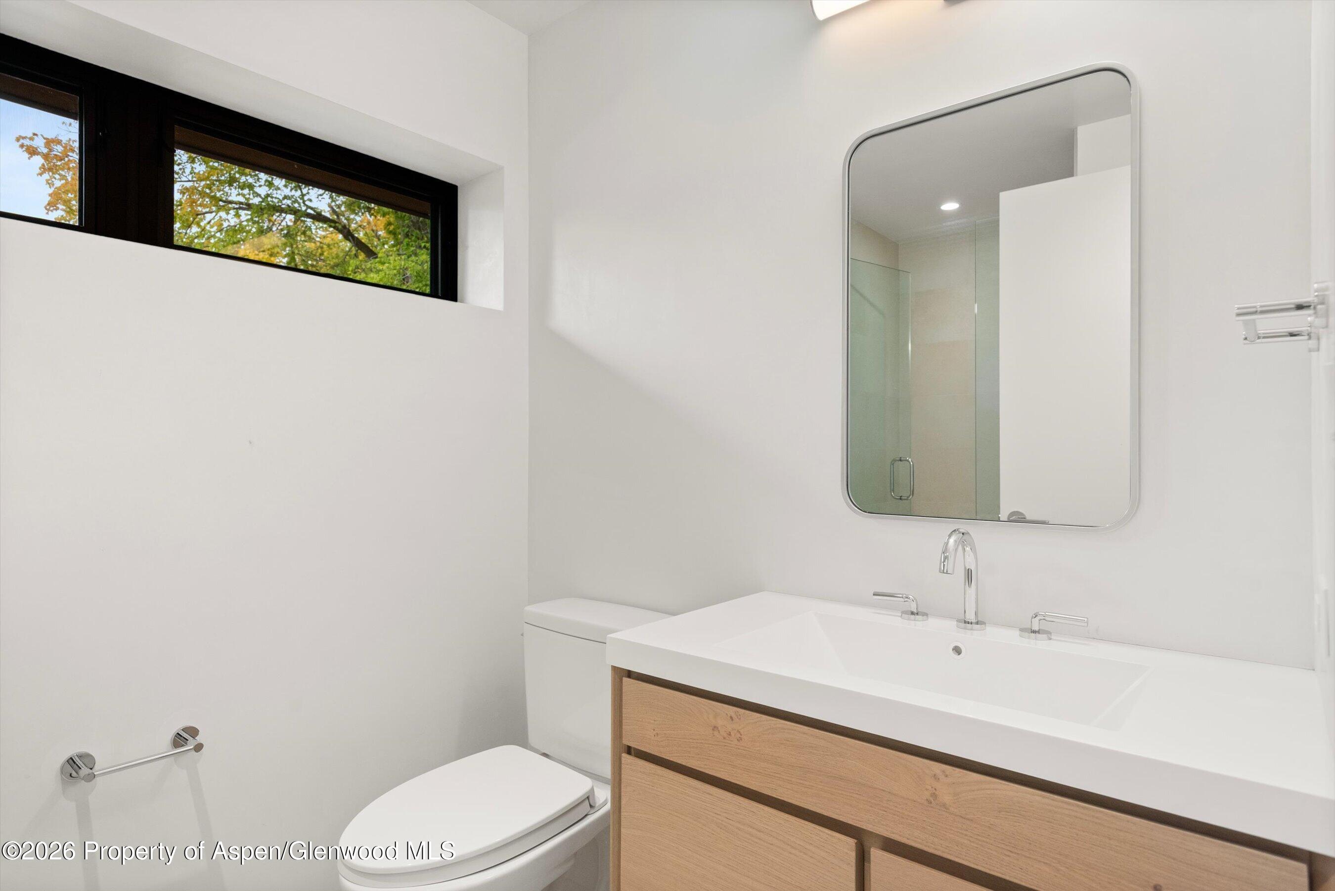 Modern bathroom with a light wood vanity, rounded mirror, and high window overlooking autumn foliage.
