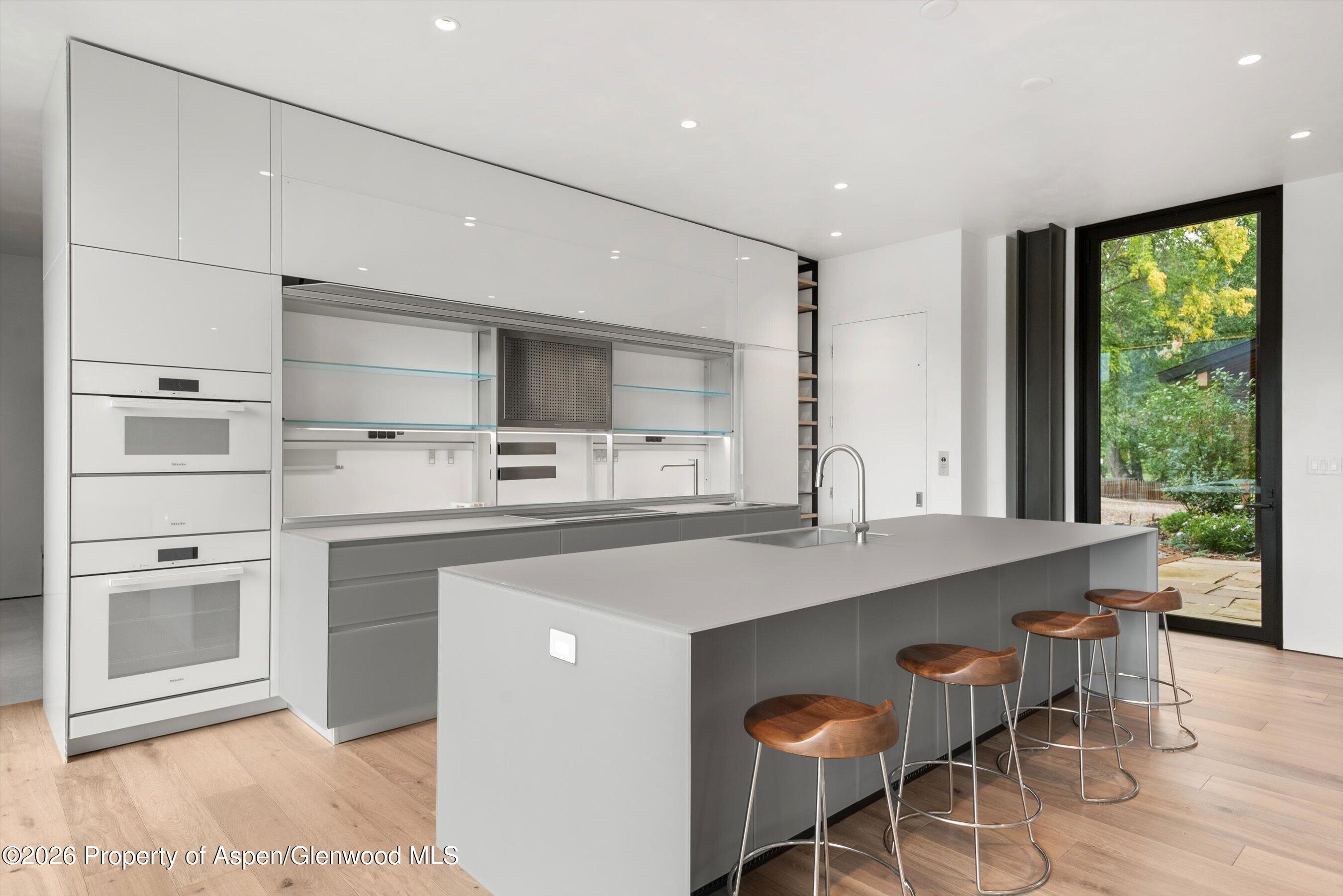 Modern kitchen with a large gray island, four wooden stools, and a black-framed glass door to the patio.