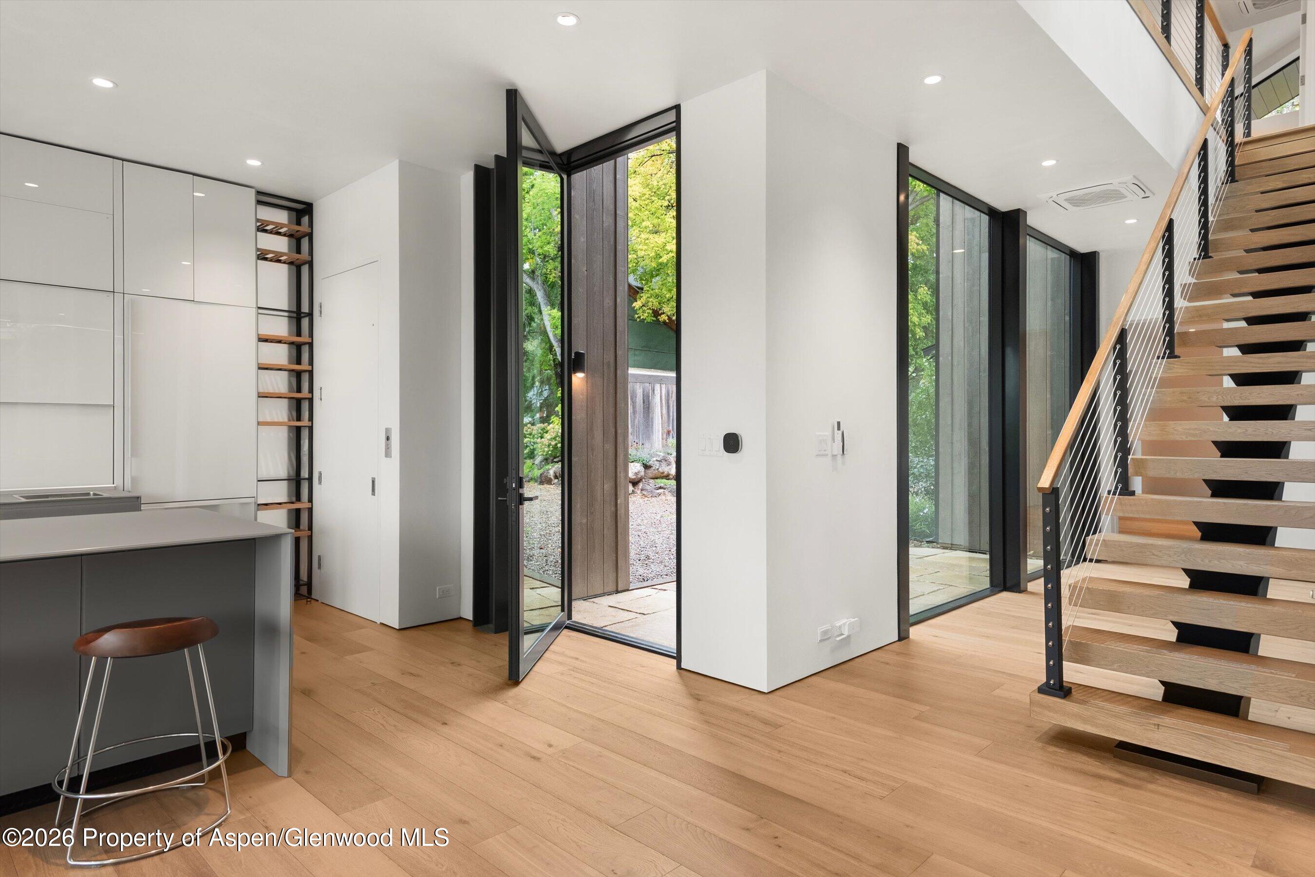 Modern entry foyer with wide-plank wood floors, glass doors, a gray kitchen island, and an open wood staircase.