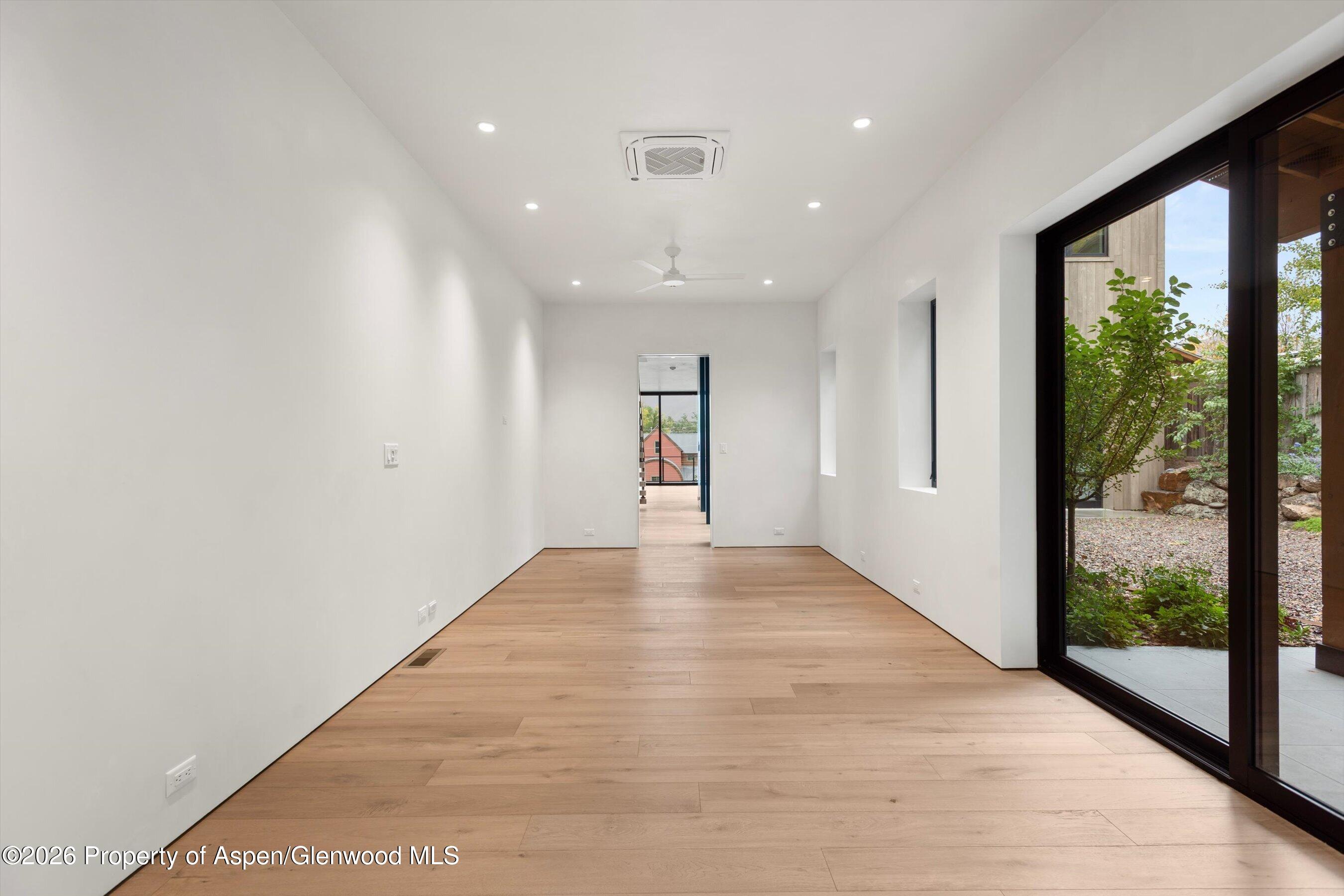 Empty modern room with pale wood floors and wide black-framed sliding glass doors overlooking a garden.