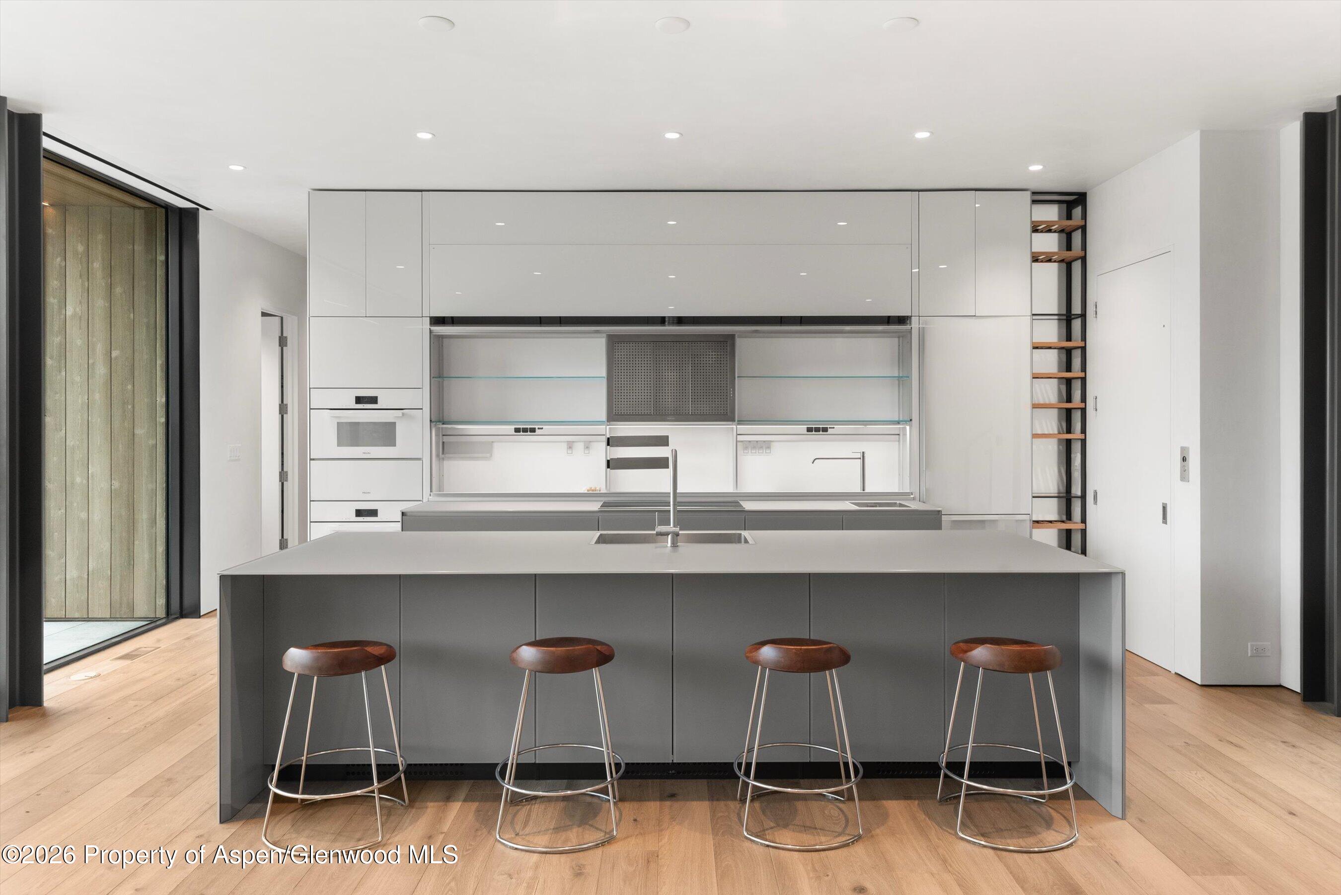 Modern kitchen with a large gray island, four brown stools, white cabinetry, and floor-to-ceiling glass doors.