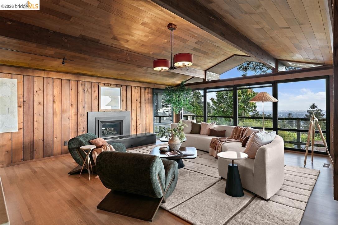Staged living room with a fireplace, wood-paneled ceiling, and broad windows overlooking trees and the city.