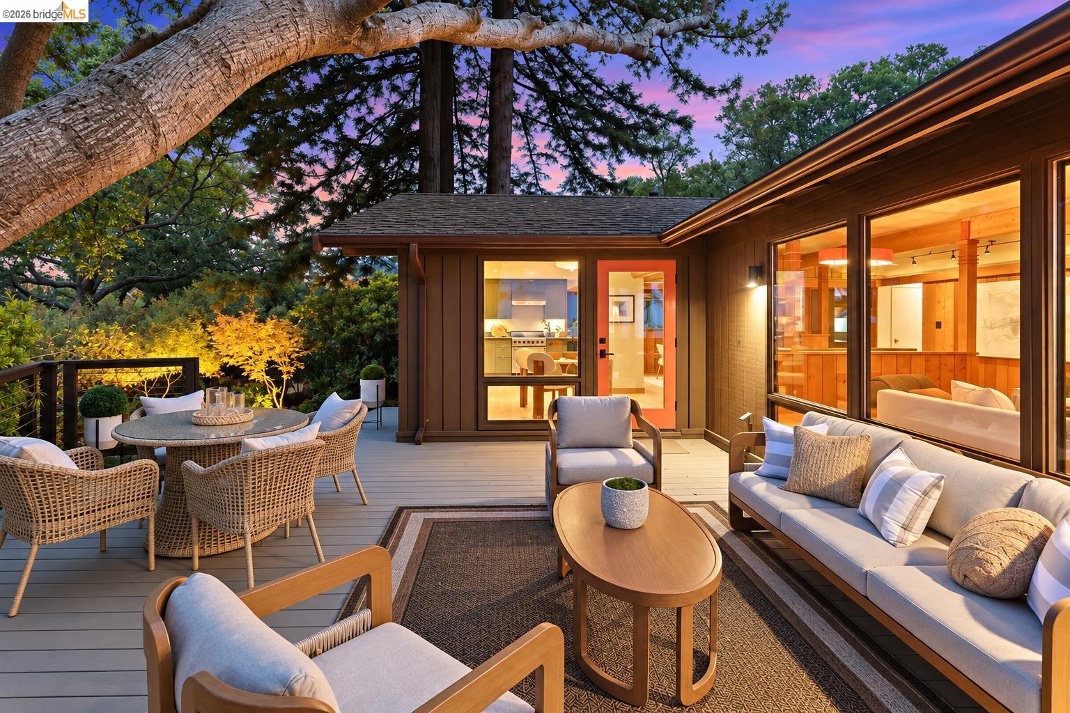 Furnished wooden deck at dusk with wicker dining set, cushioned lounge seating, and warm-lit home windows.