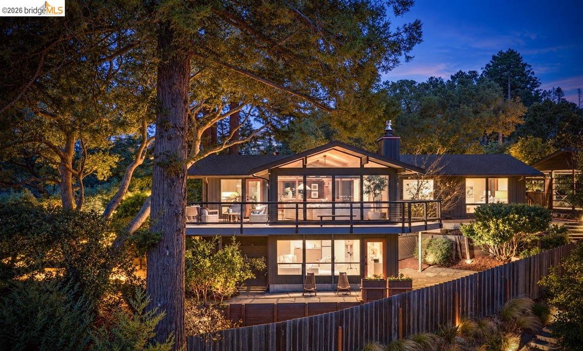 Modern home with a broad glass-fronted deck, warmly lit at dusk among mature trees.