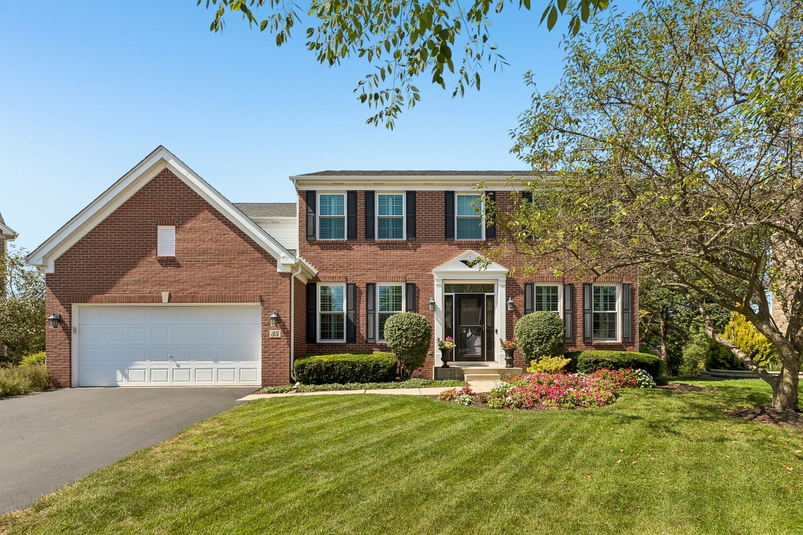Two-story red-brick house with black shutters, a white garage door, and landscaped flower beds.