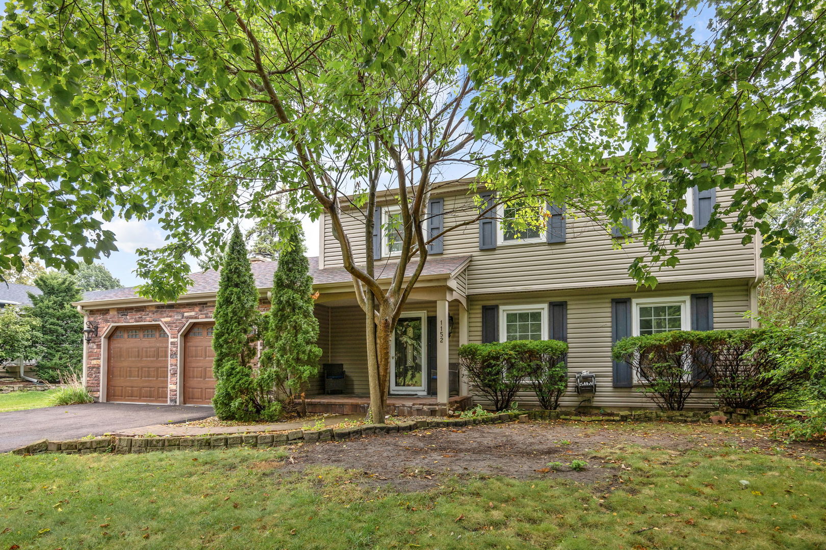 Two-story beige house with a stone-faced garage, dark shutters, and a leafy tree shading the front yard.