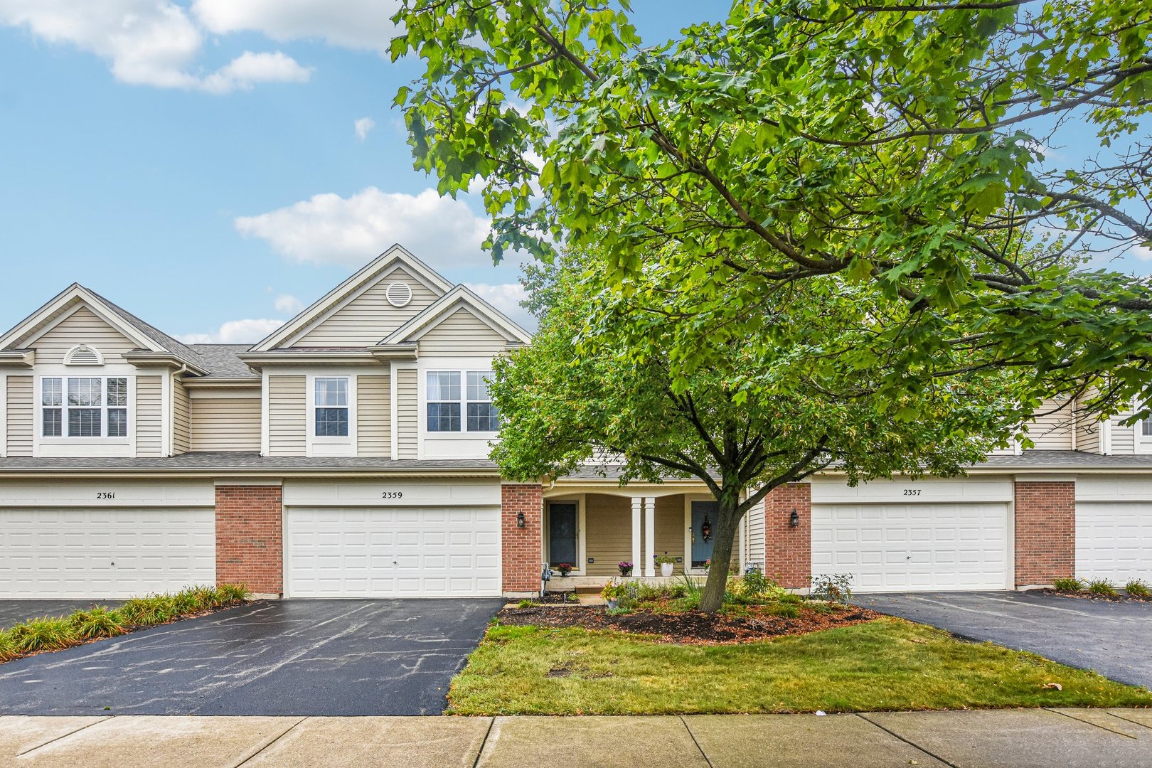 Attached homes with beige siding and brick accents, shaded by a large leafy tree.