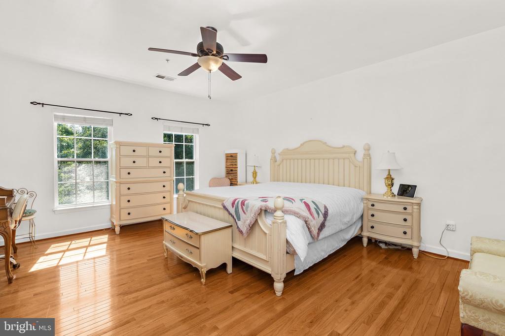 Furnished bedroom with a cream bed, matching dressers, hardwood floors, and sunlight through two windows.