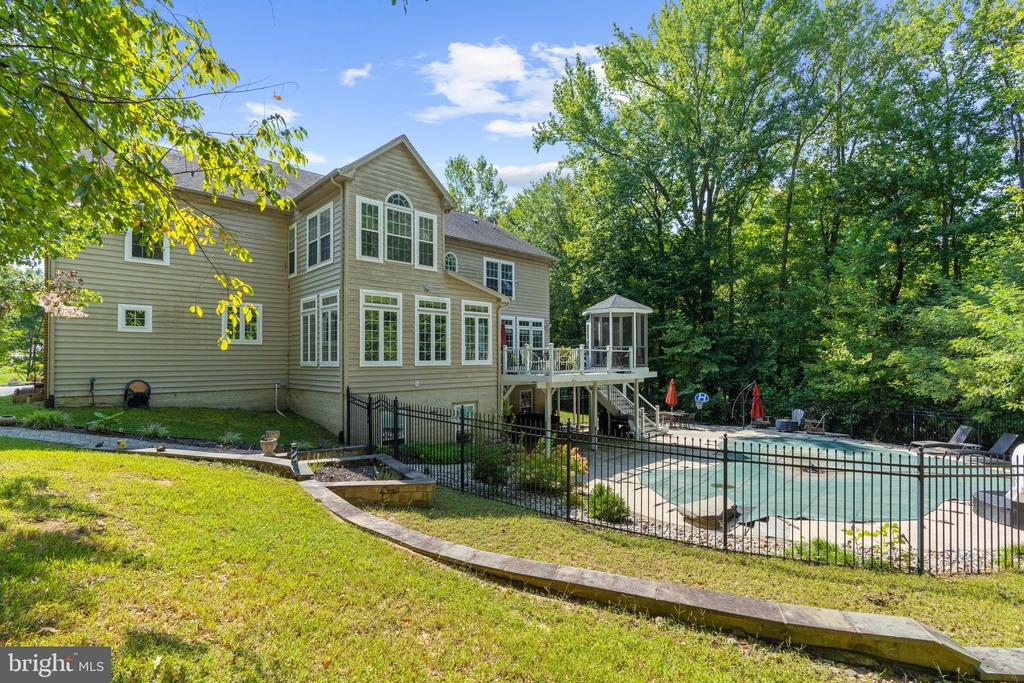Rear of a beige two-story home with a raised deck and fenced pool among mature trees.