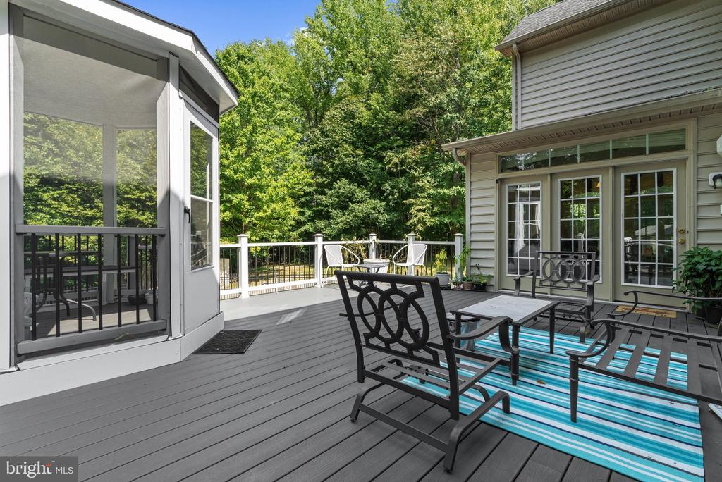 Composite deck with black patio furniture and blue striped rug beside a screened porch, overlooking trees.