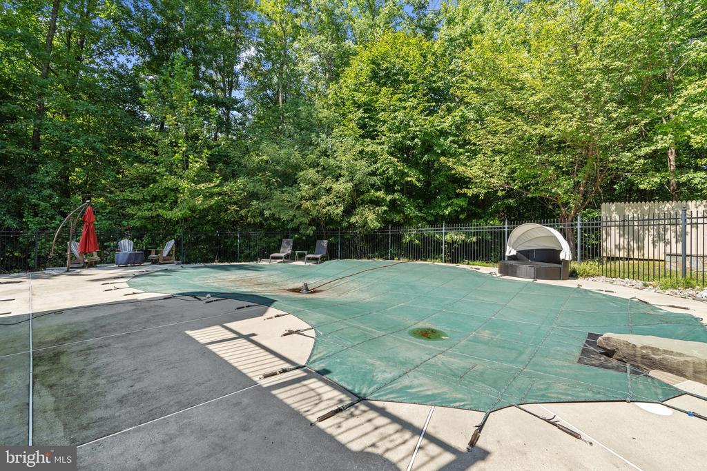 Fenced outdoor pool covered with a green cover, surrounded by a concrete deck and wooded greenery.