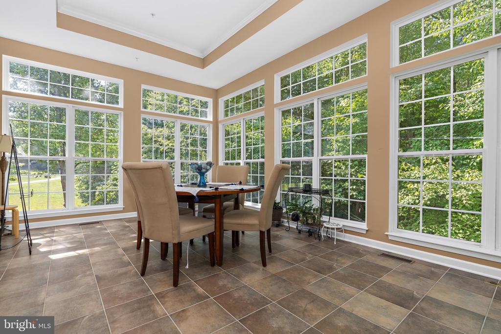Sunroom with a round dining table, tiled floor, and large windows overlooking leafy greenery.