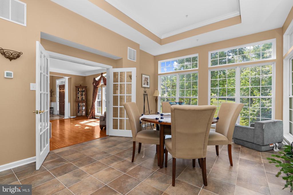 Dining area with tall windows, tiled flooring, and French doors opening to a hardwood-floored room.