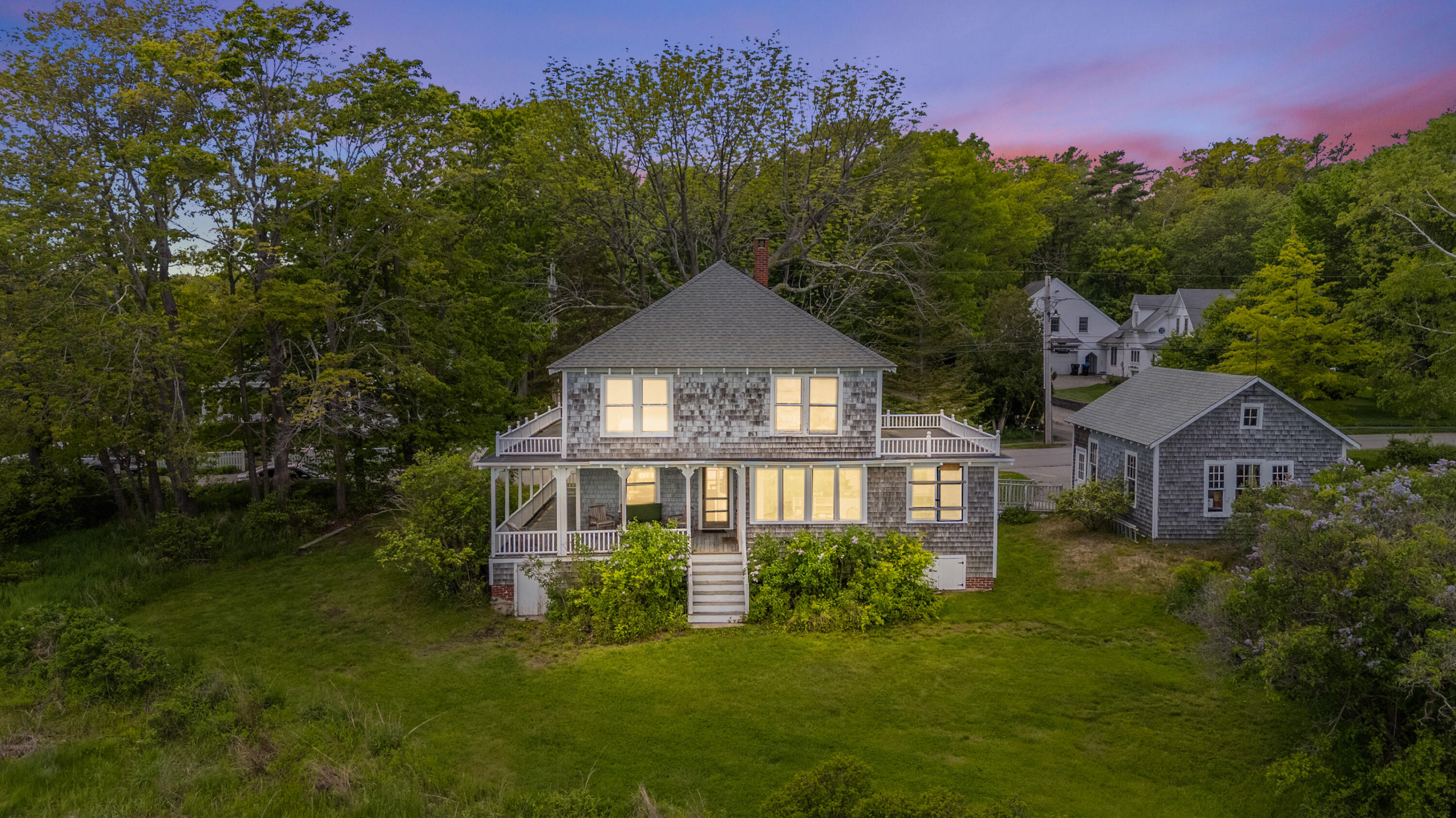 Gray-shingled home with wraparound porch and detached outbuilding, surrounded by trees at dusk.